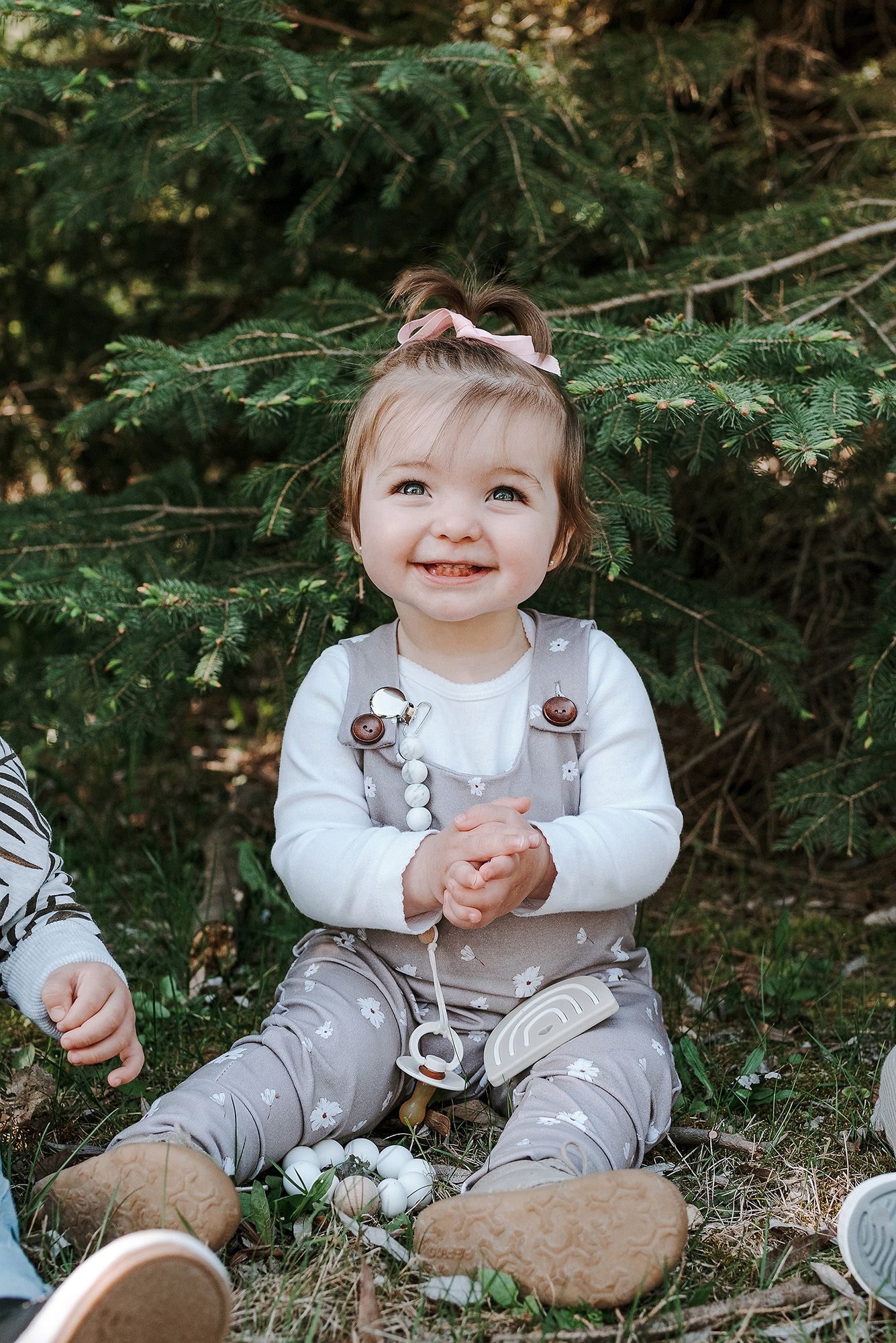 A toddler in gray overalls and white shirt sits happily on the ground near evergreen branches.