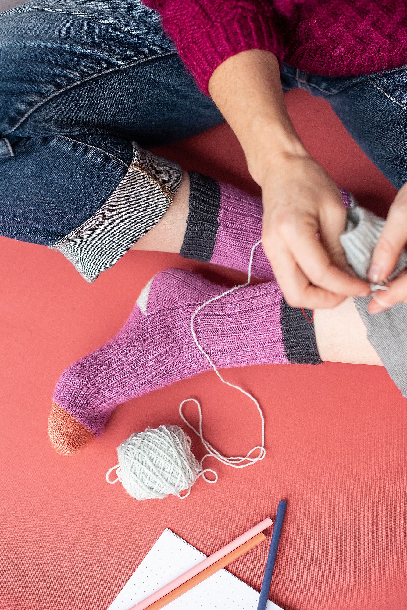 Hands darning pink knitted sock with white yarn and needle.
