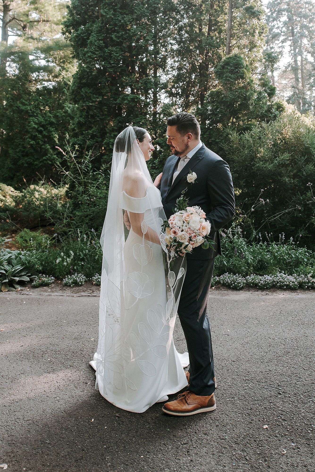 A romantic wedding couple shares an intimate moment on a gravel path surrounded by lush garden greenery.