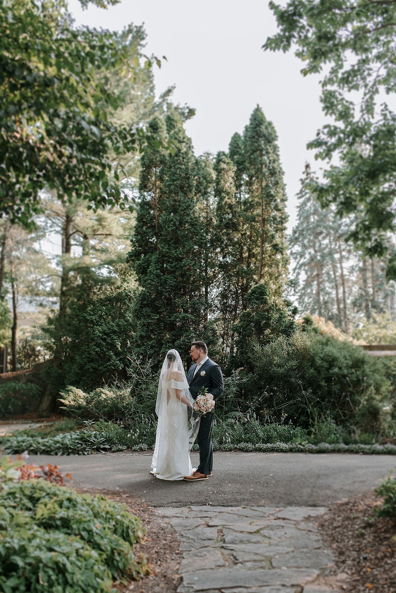 A couple shares an intimate moment on a stone pathway surrounded by lush greenery in an outdoor garden setting.