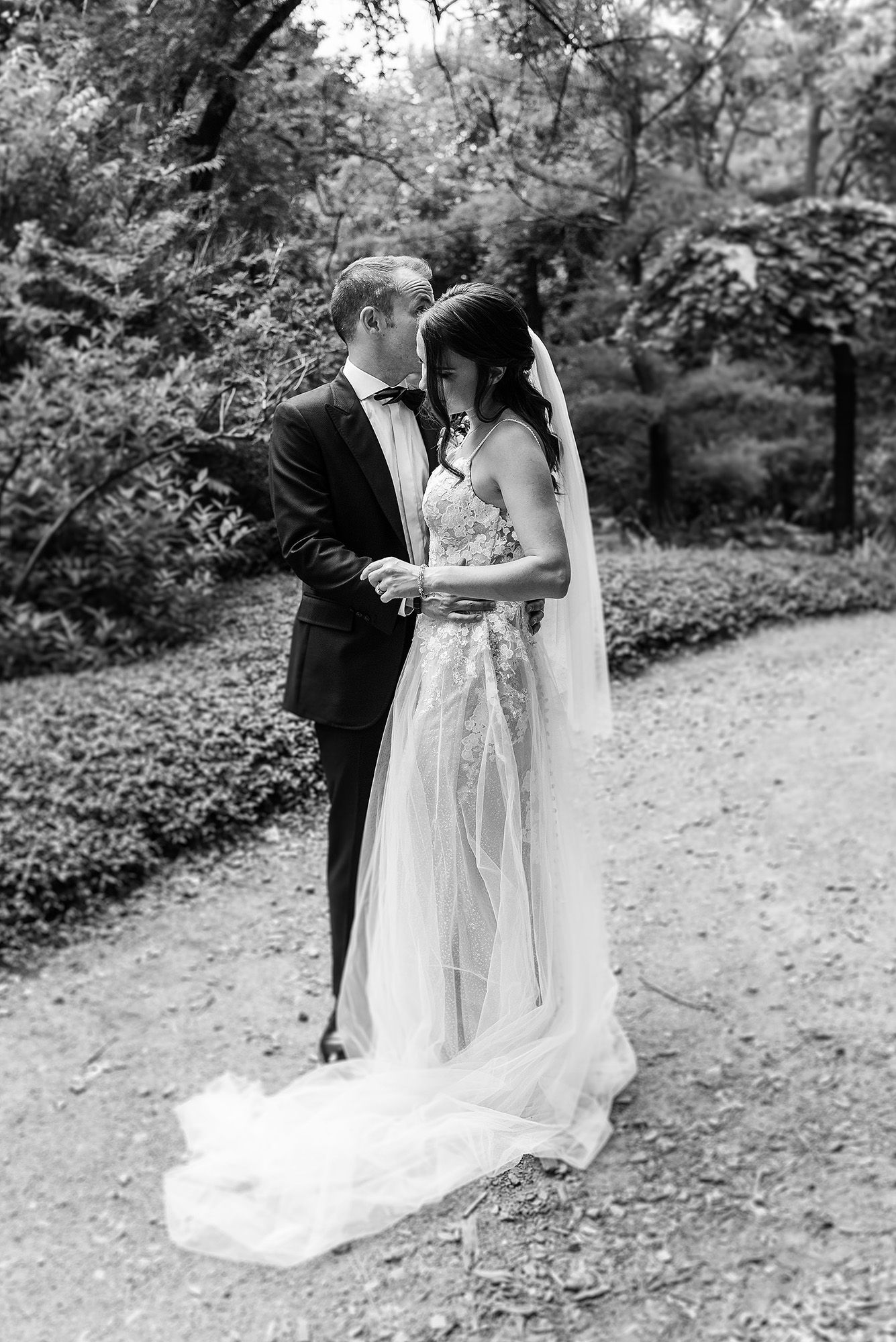 Black and white photo of newlyweds embracing on garden path with flowing wedding dress and veil.