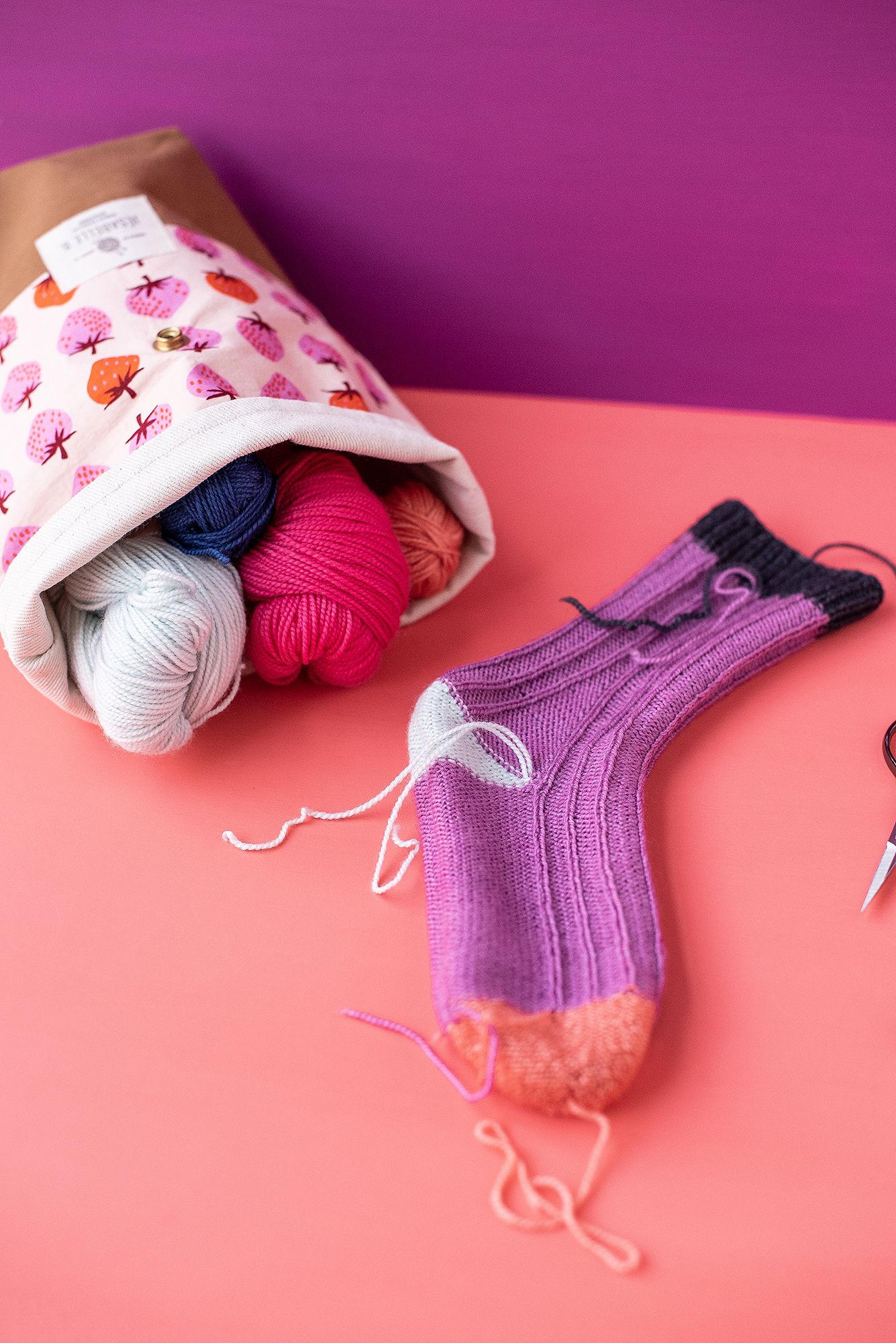 Knitting project with purple sock and yarn balls spilling from floral project bag.