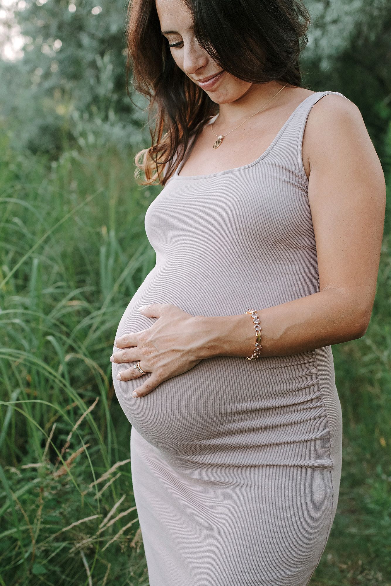 Woman in gray tank top cradling her pregnant belly outdoors among tall grass.