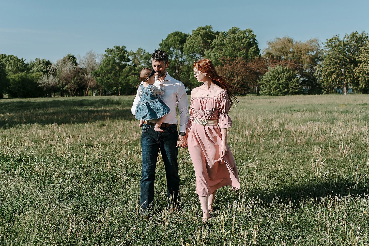A couple walks hand in hand through a sunlit meadow wearing casual attire on a warm afternoon.