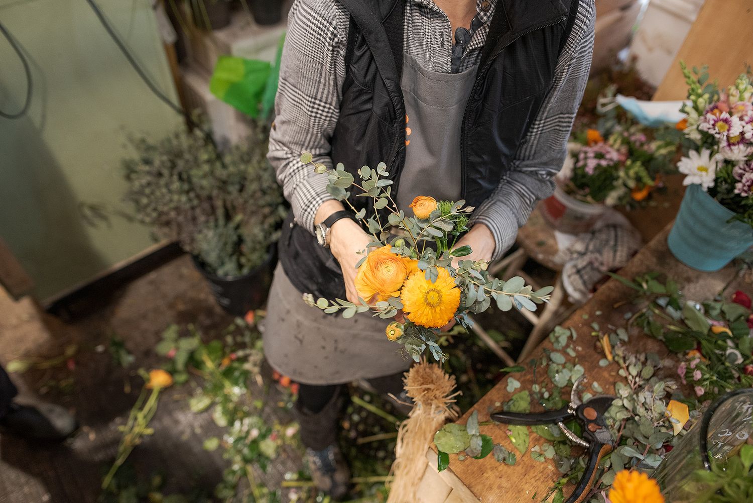 A florist arranges fresh flowers and greenery in a rustic outdoor setting with natural lighting.