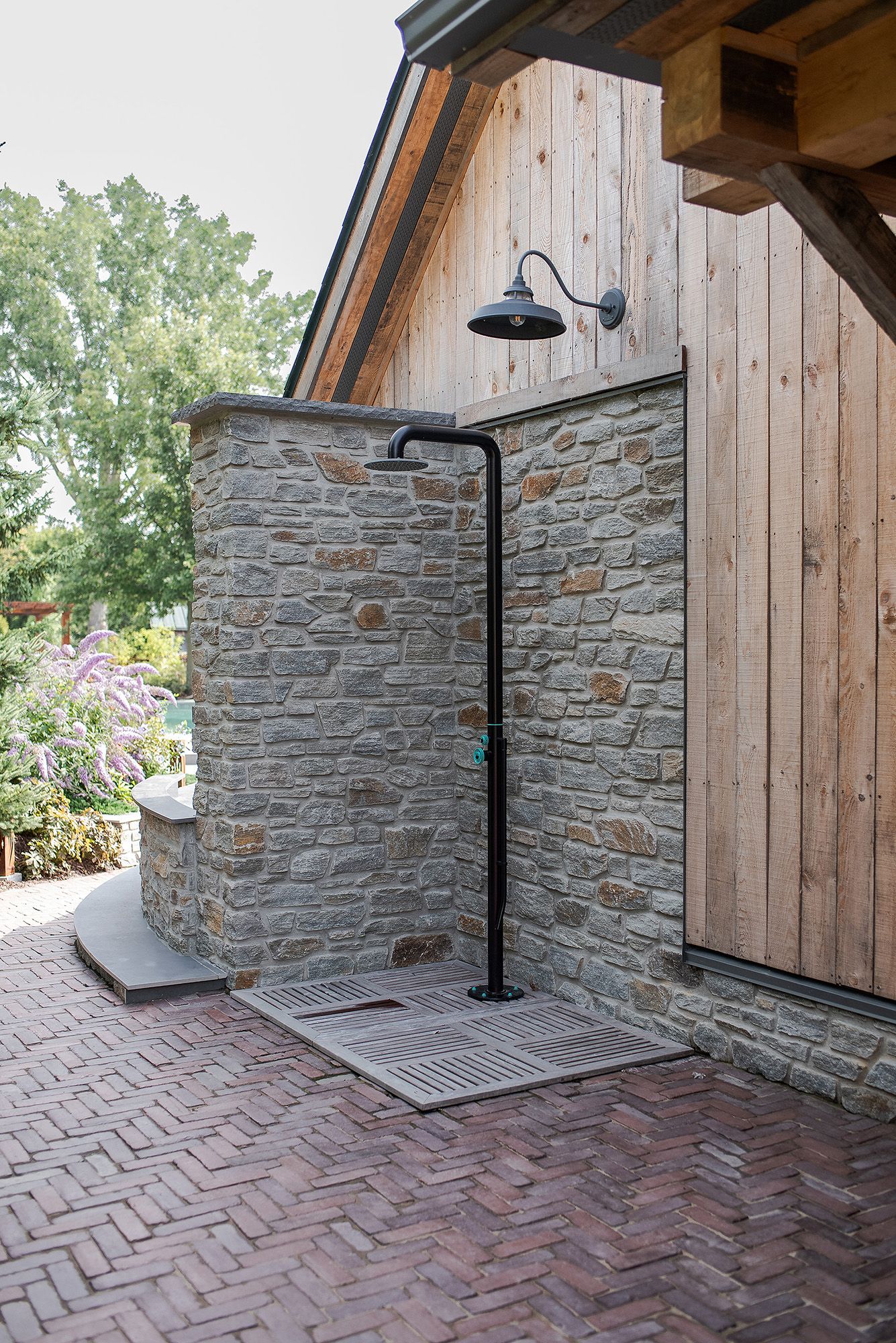 Herringbone brick patio meets rustic stone wall and wooden siding with black outdoor light fixture mounted above.