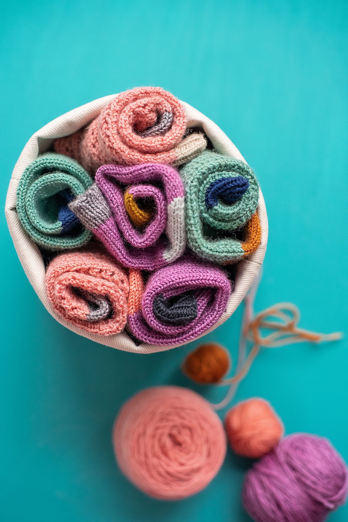 Rolled up colorful knitted socks arranged in white bowl against turquoise background.