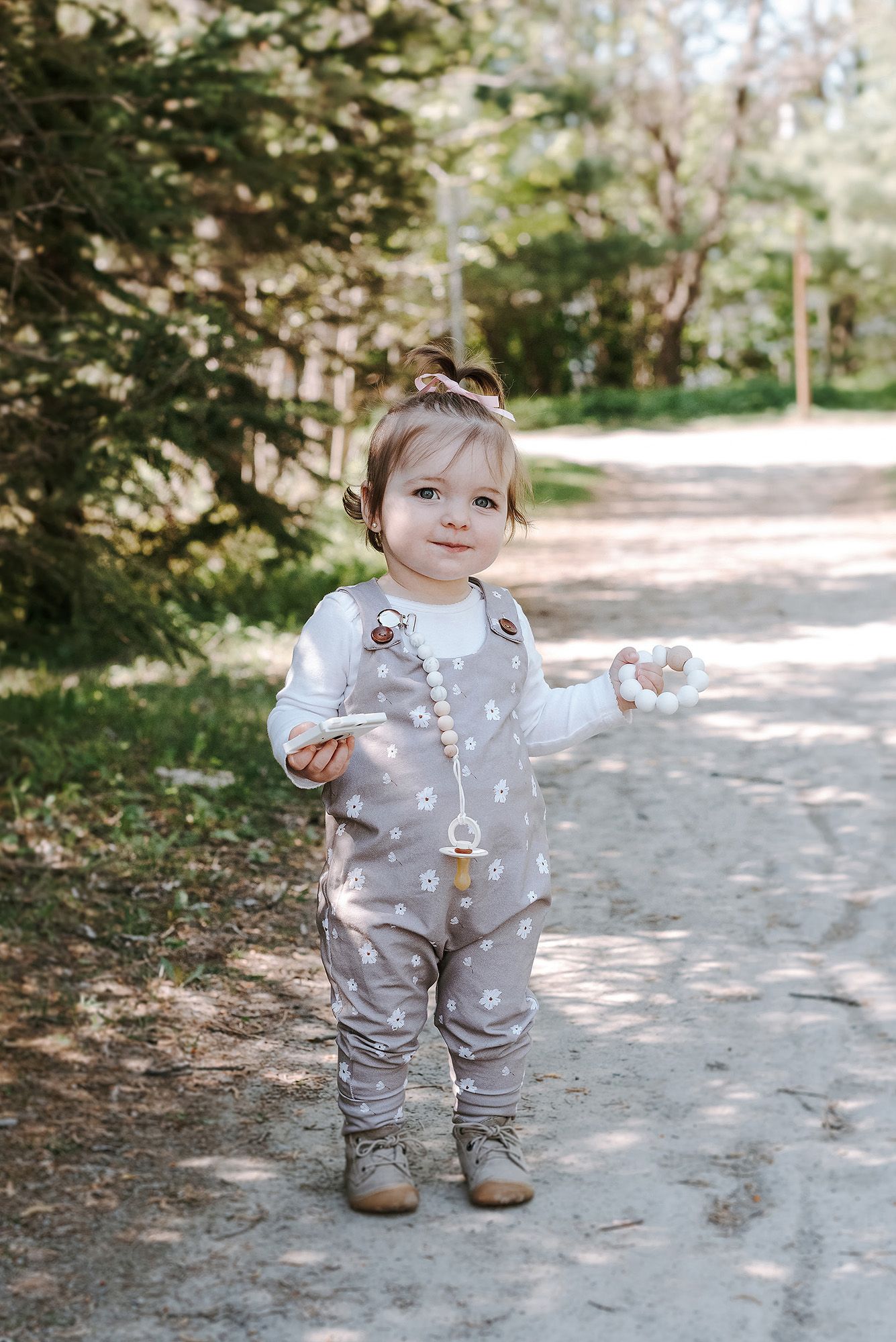 Baby in gray patterned outfit standing on a path surrounded by trees.