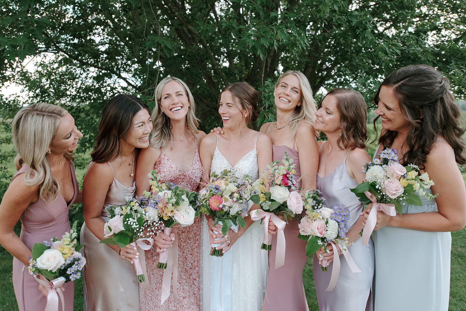 A wedding party in pink and mauve dresses poses together outdoors while holding floral bouquets.