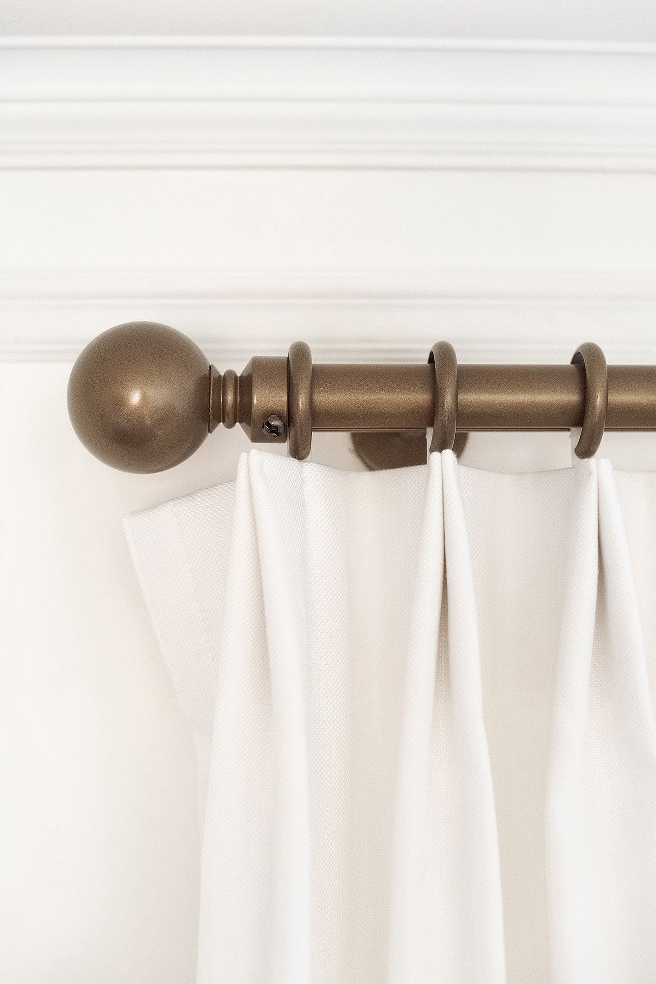 White curtain panels hanging from a bronze curtain rod with decorative finials against a light wall.