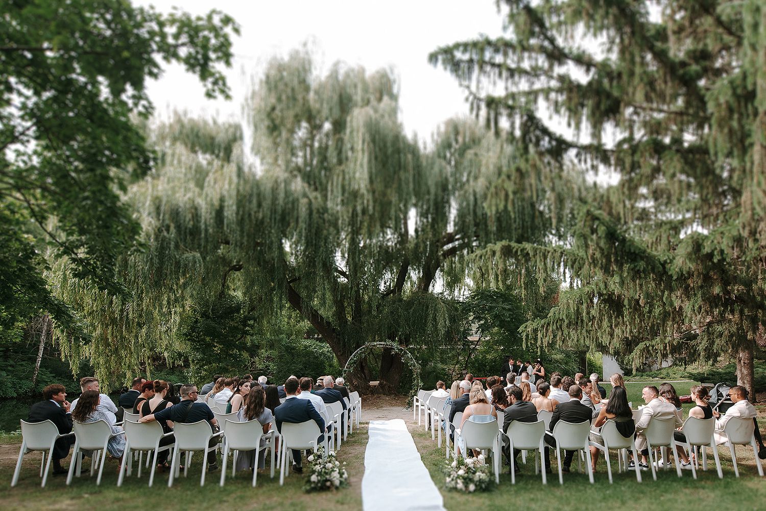A romantic outdoor wedding ceremony setup under weeping willow trees with white chairs and floral arrangements.
