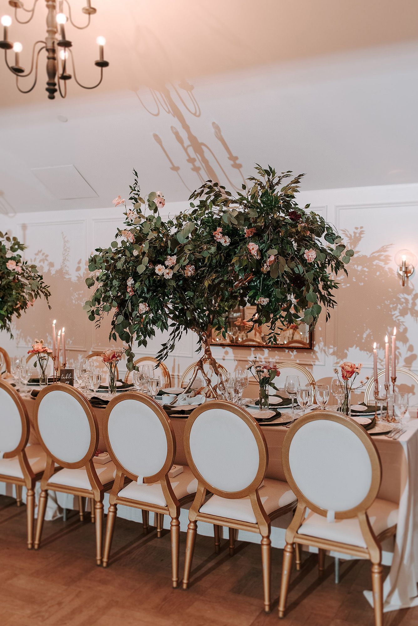 Elegant wedding reception table setup with white upholstered chairs and lush greenery centerpieces under chandeliers.