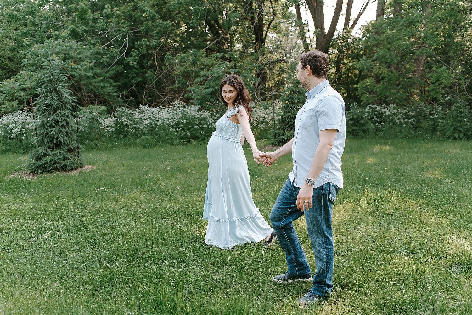 A romantic couple in white and denim outfits poses together in a lush green outdoor setting during a photo session.