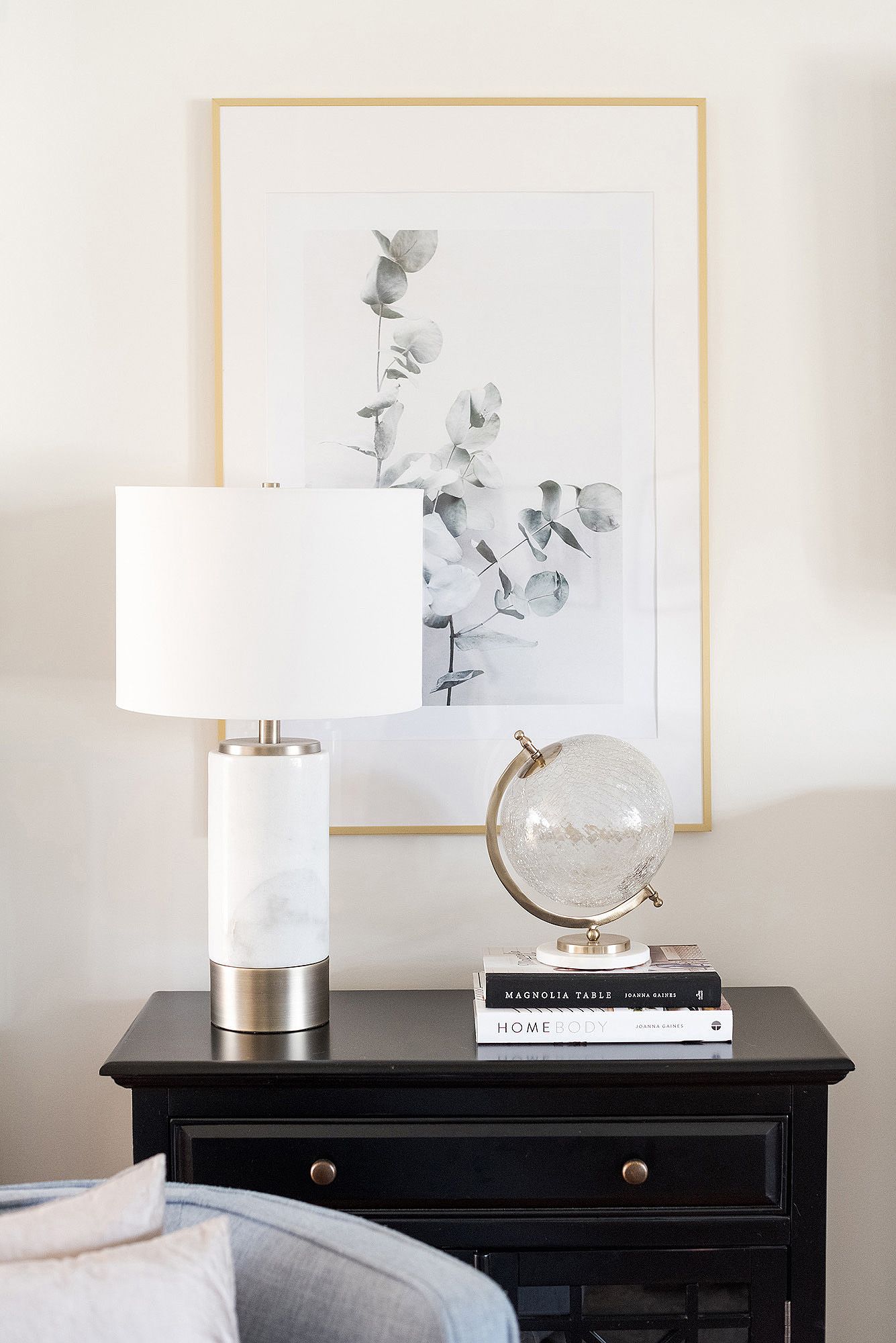 A black nightstand with a white table lamp, glass globe, and framed botanical artwork creates an elegant bedroom vignette.