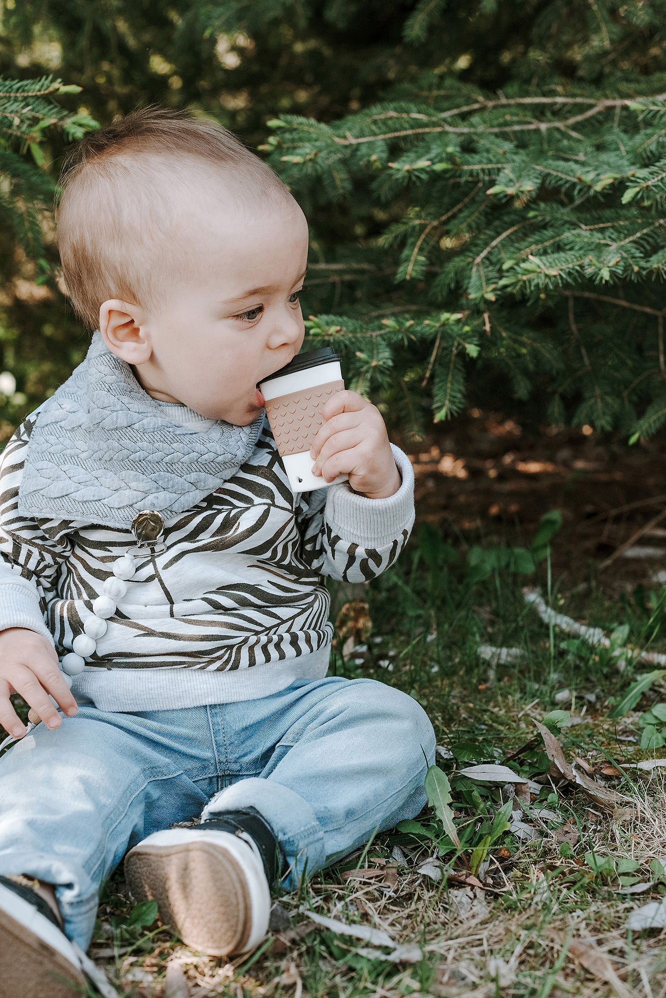 A baby in a patterned sweater and jeans sits under evergreen branches drinking from a cup.