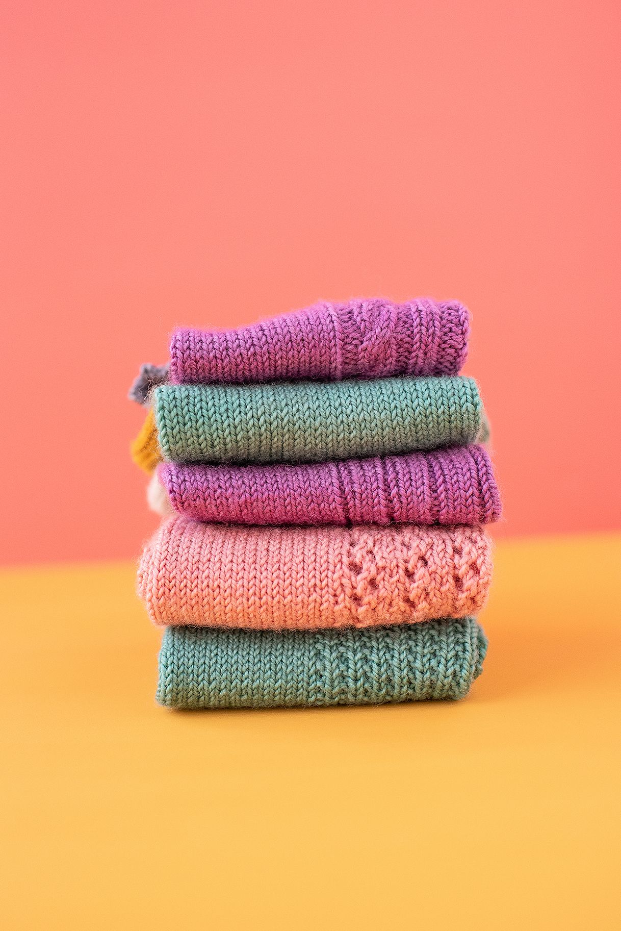 Neatly folded stack of colorful knitted socks on pink and orange background.