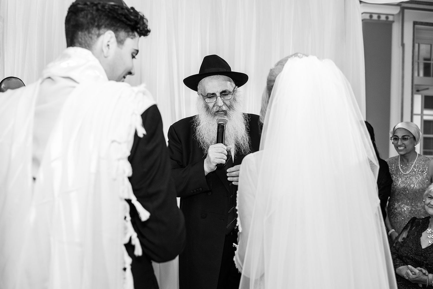 Rabbi officiating a Jewish wedding ceremony with bride and groom under the chuppah in black and white.