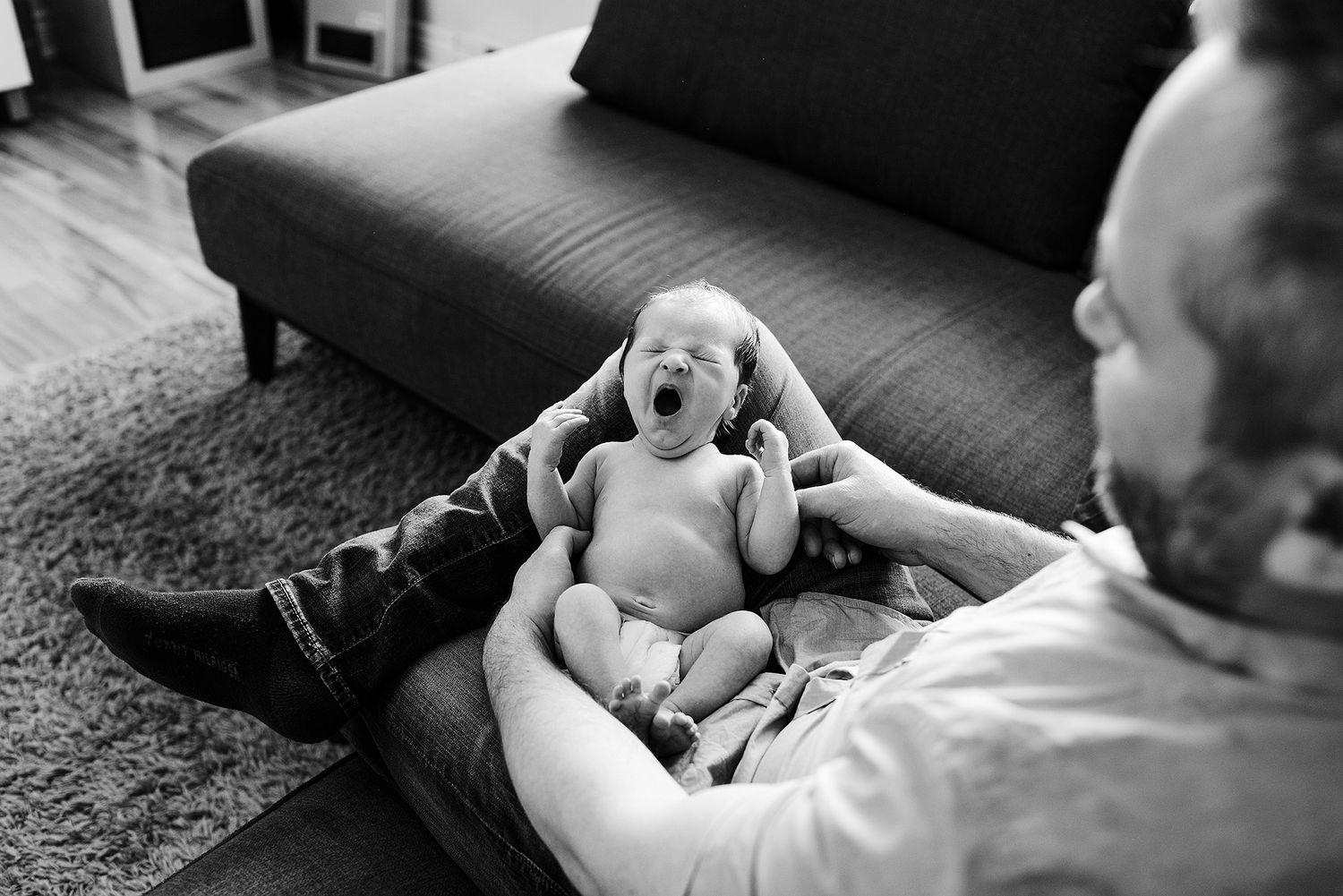 Parent tenderly cradling newborn on dark gray couch in moody lighting.