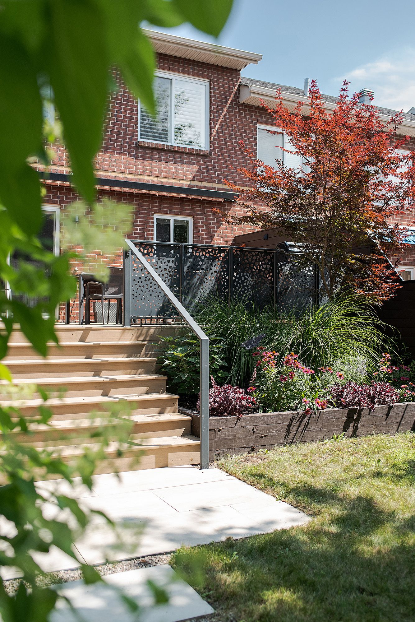 Modern wooden deck with glass railing extends from brick house into landscaped backyard with ornamental grasses and flowers.