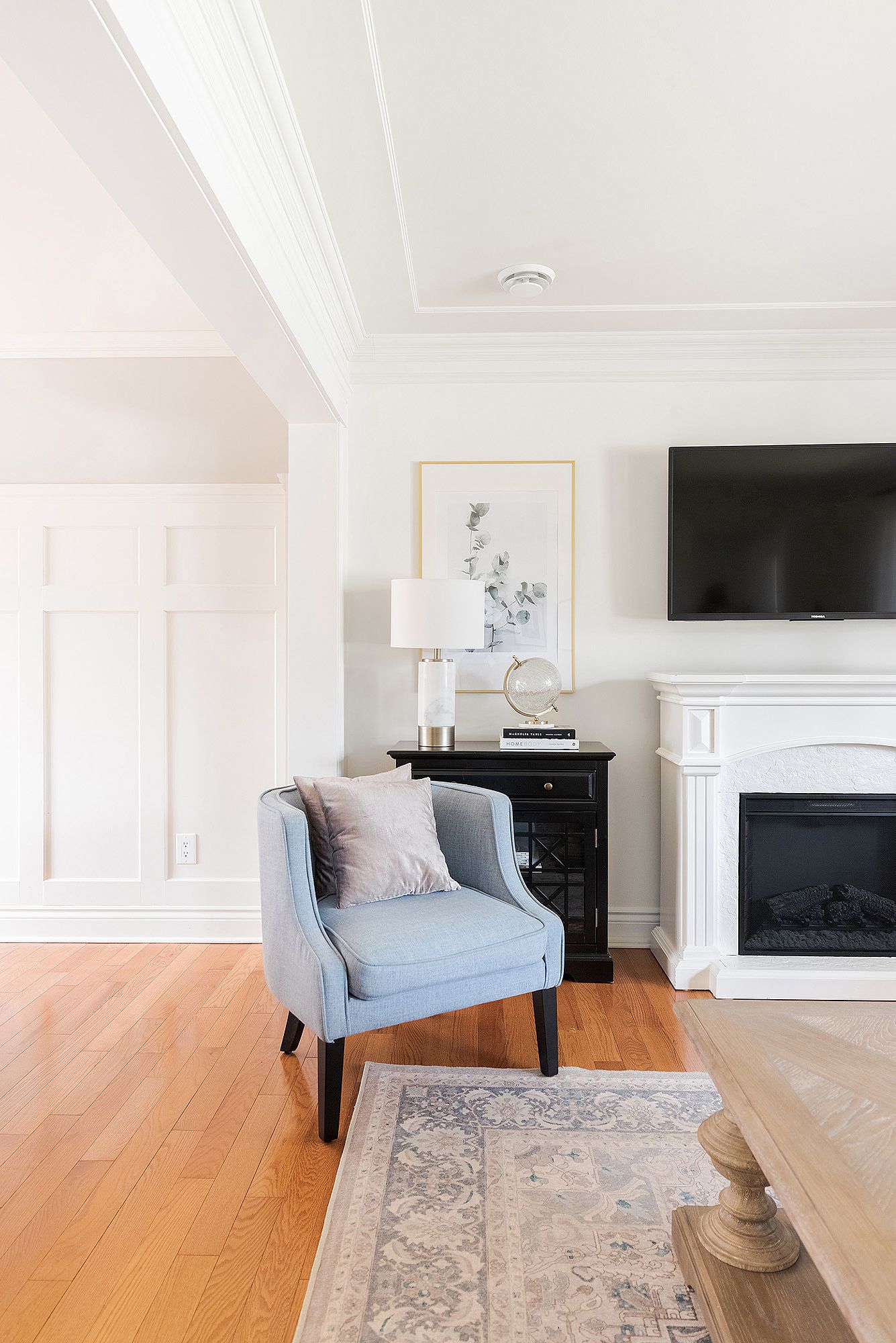 Modern living room with light blue armchair, hardwood floors, and area rug near white fireplace and mounted TV.