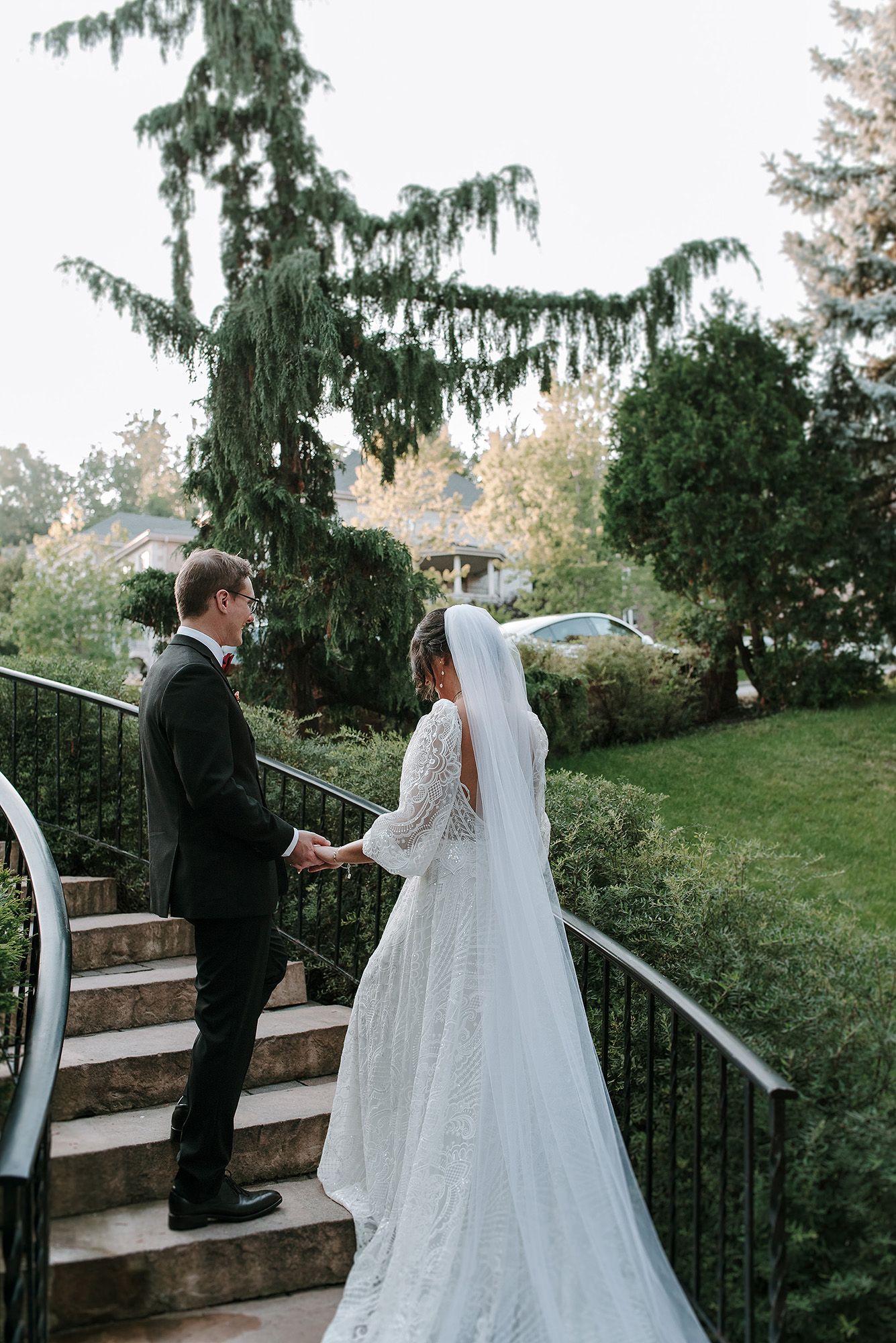 A couple in formal wedding attire shares a private moment on outdoor stairs surrounded by evergreen trees.