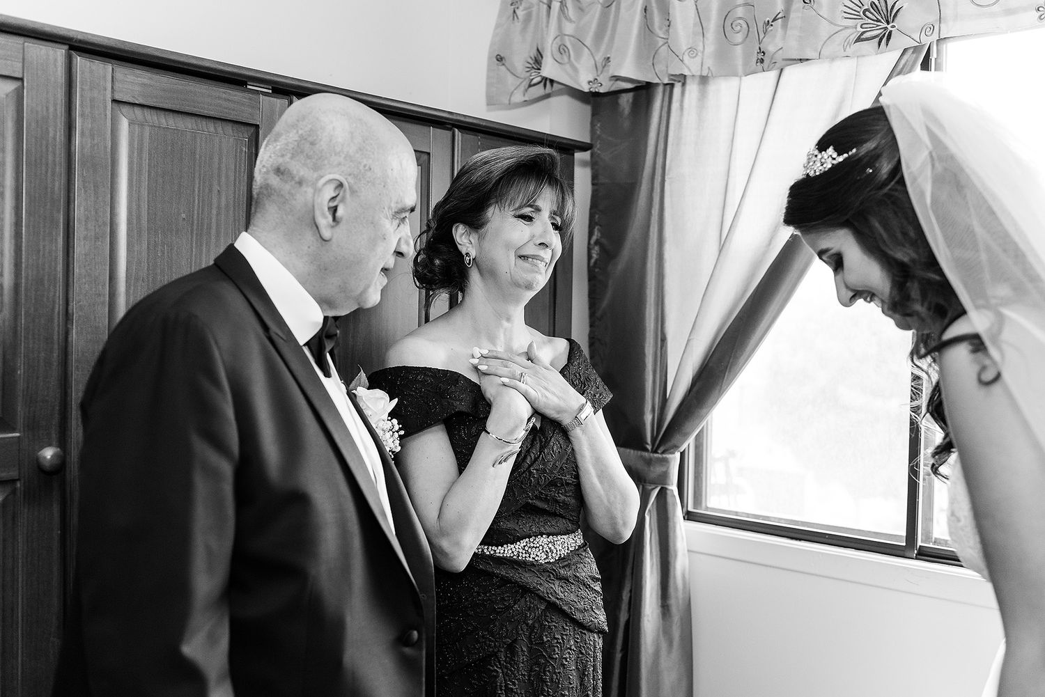 Three people in formal attire share a candid moment near a window in black and white.