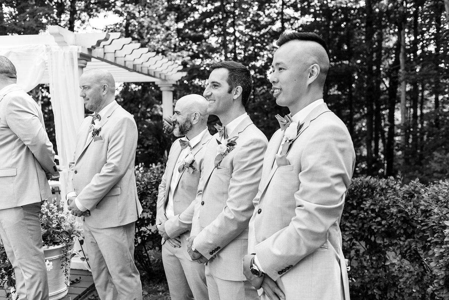 A group of groomsmen in light colored suits stand together during an outdoor wedding ceremony under a pergola.