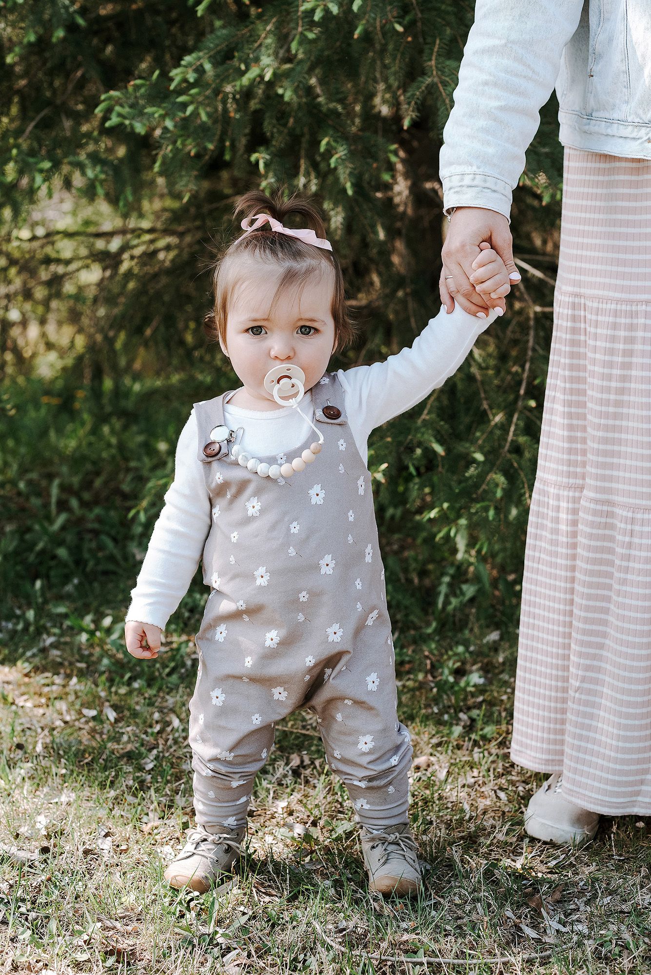A toddler in floral overalls and white shirt holds hands with someone while standing outdoors.