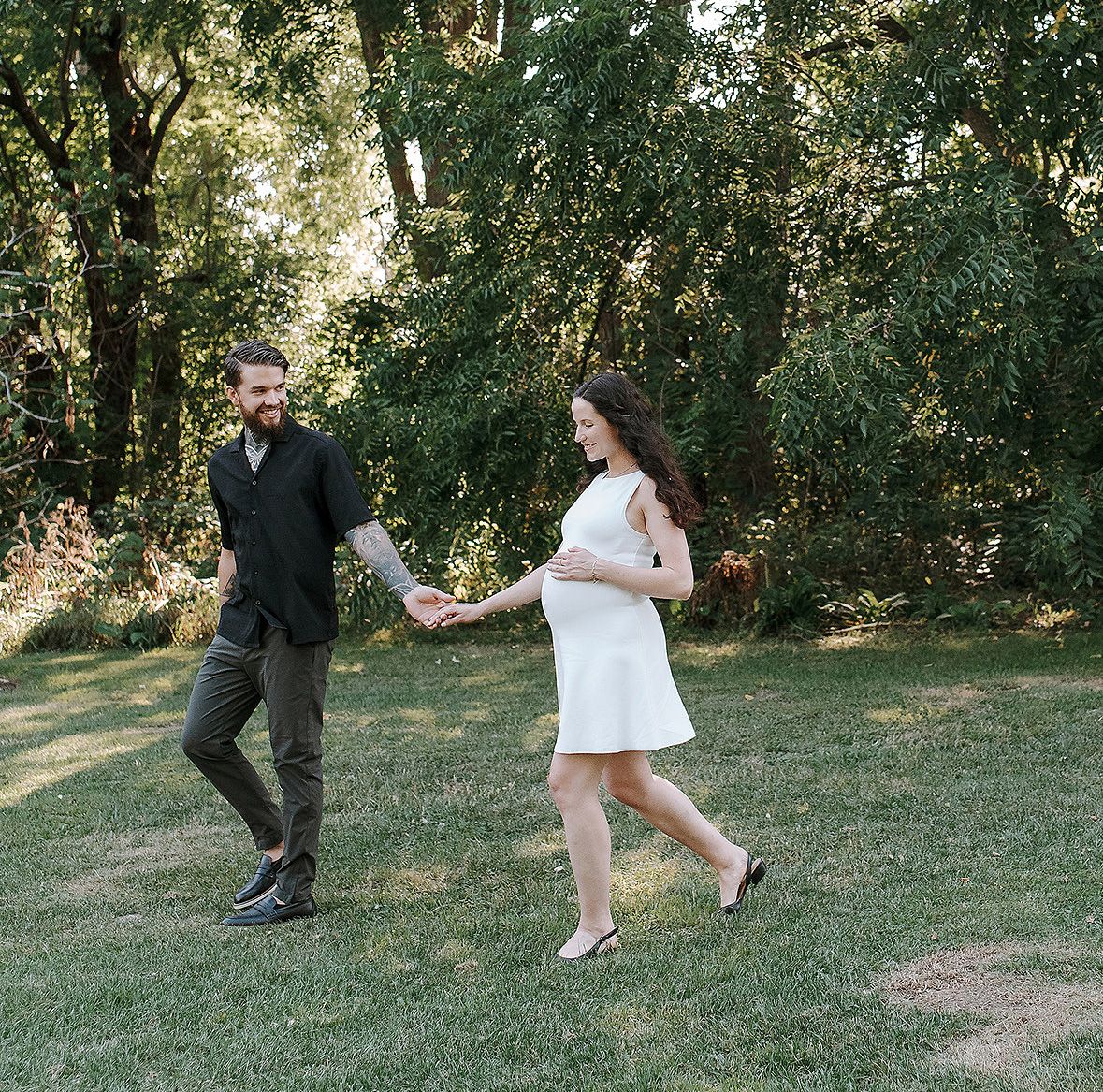 A couple holds hands while walking together in a sunlit wooded area on a summer day.
