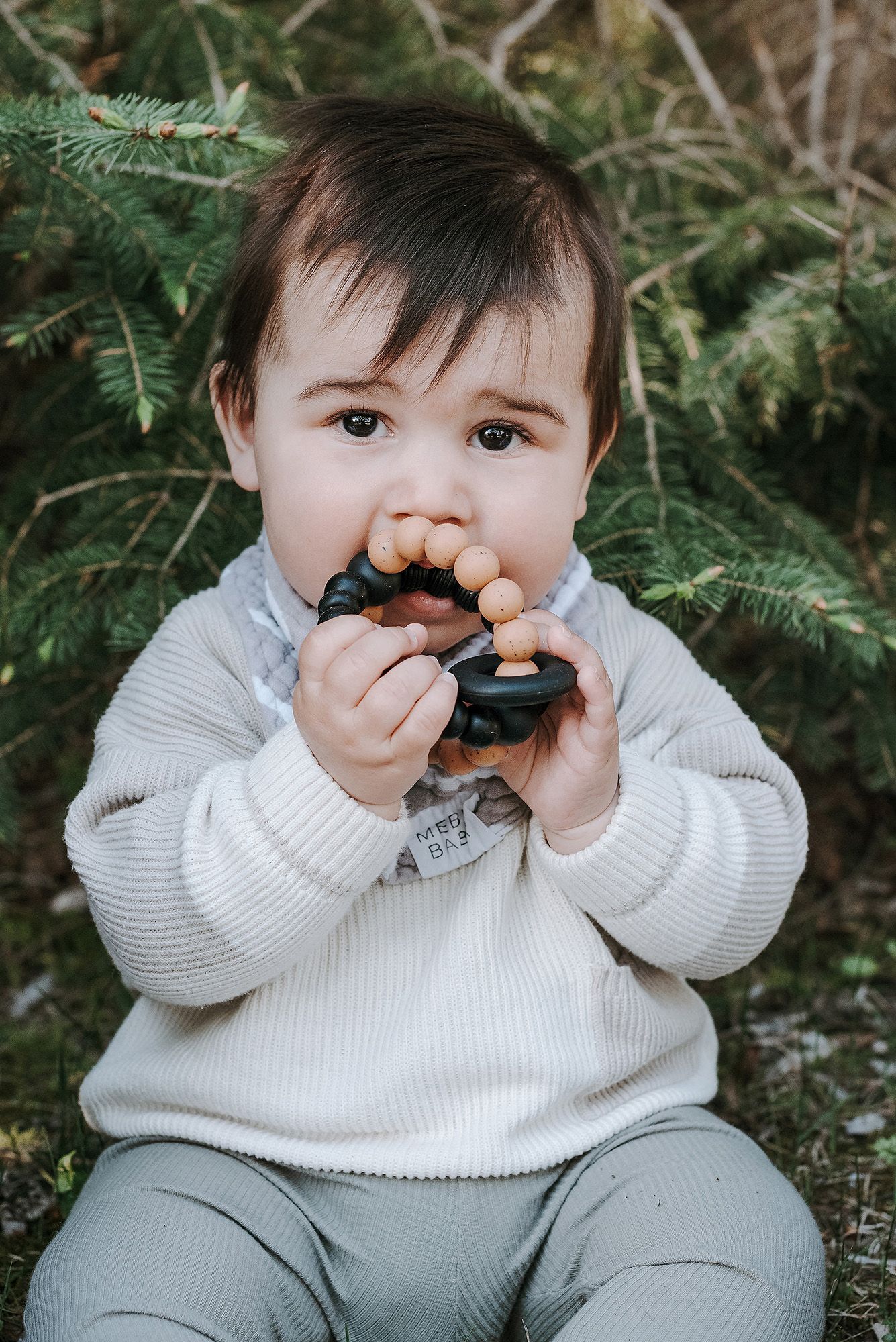 Baby in white sweater and gray pants playing with pacifier while sitting on ground.