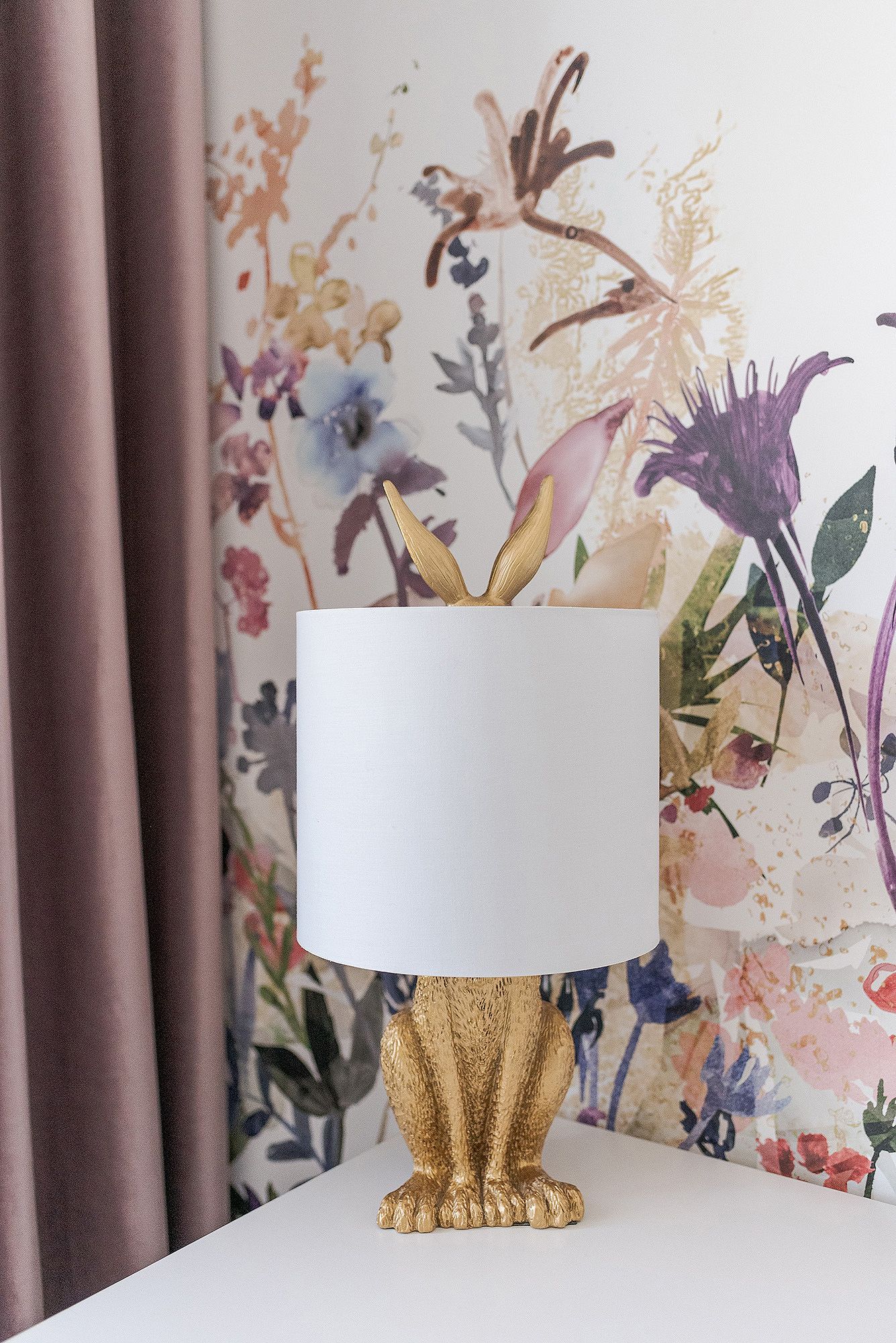 Modern white table lamp with gold base sits against floral wallpaper with pink, purple and blue blooms.