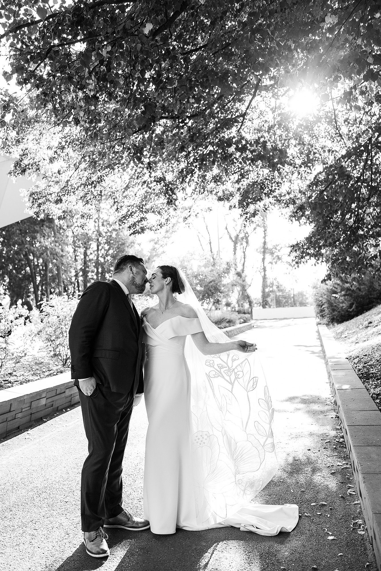 A romantic black and white wedding photo of a couple sharing an intimate moment on a sunlit path surrounded by trees.