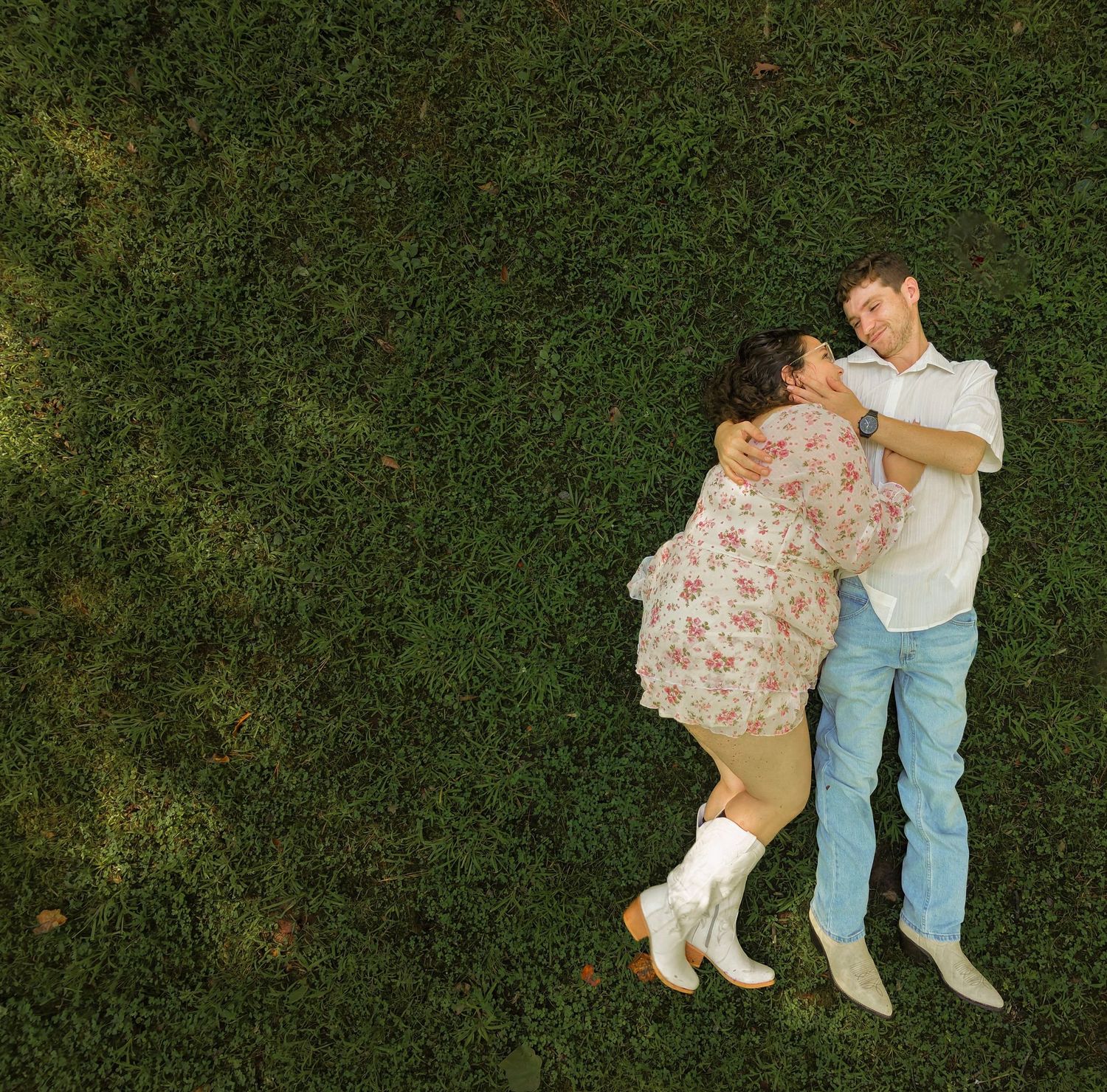 Aerial view of a couple lying on green grass, embracing romantically, woman in floral dress with white boots.