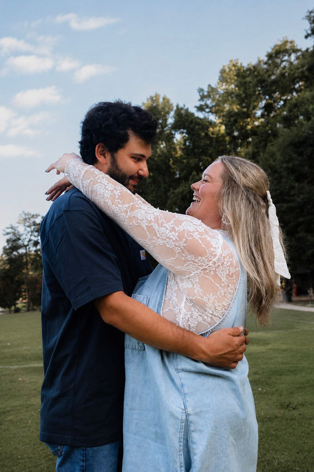 A couple embraces and smiles at each other outdoors, woman in lace top and denim skirt, man in navy shirt.