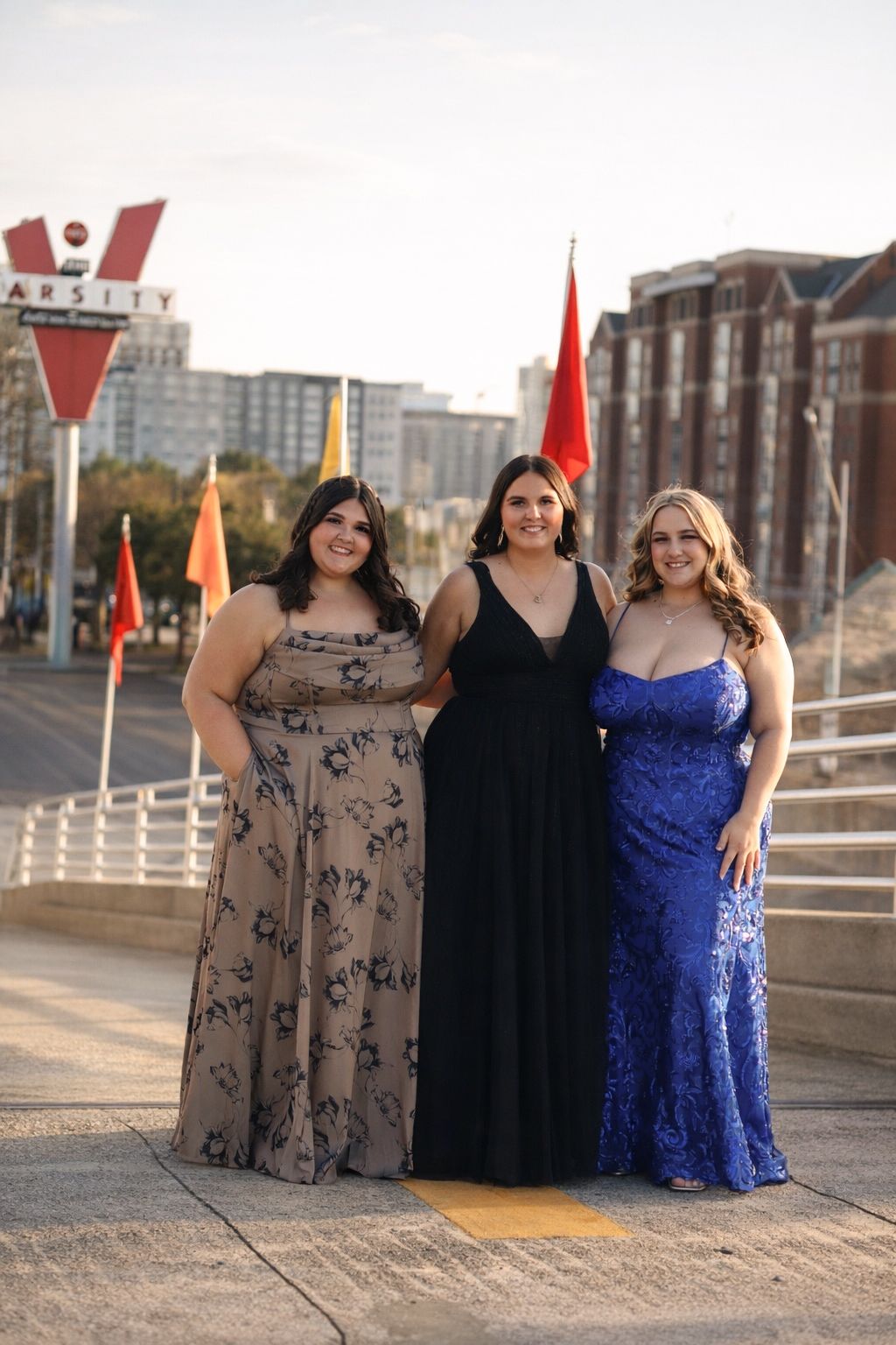 Three people in formal evening gowns pose together on a waterfront boardwalk at sunset.