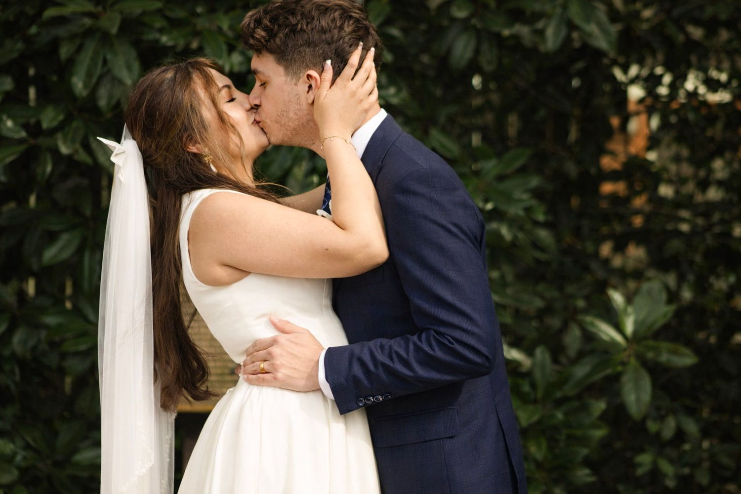 Romantic wedding couple sharing intimate kiss in garden setting with lush greenery backdrop.