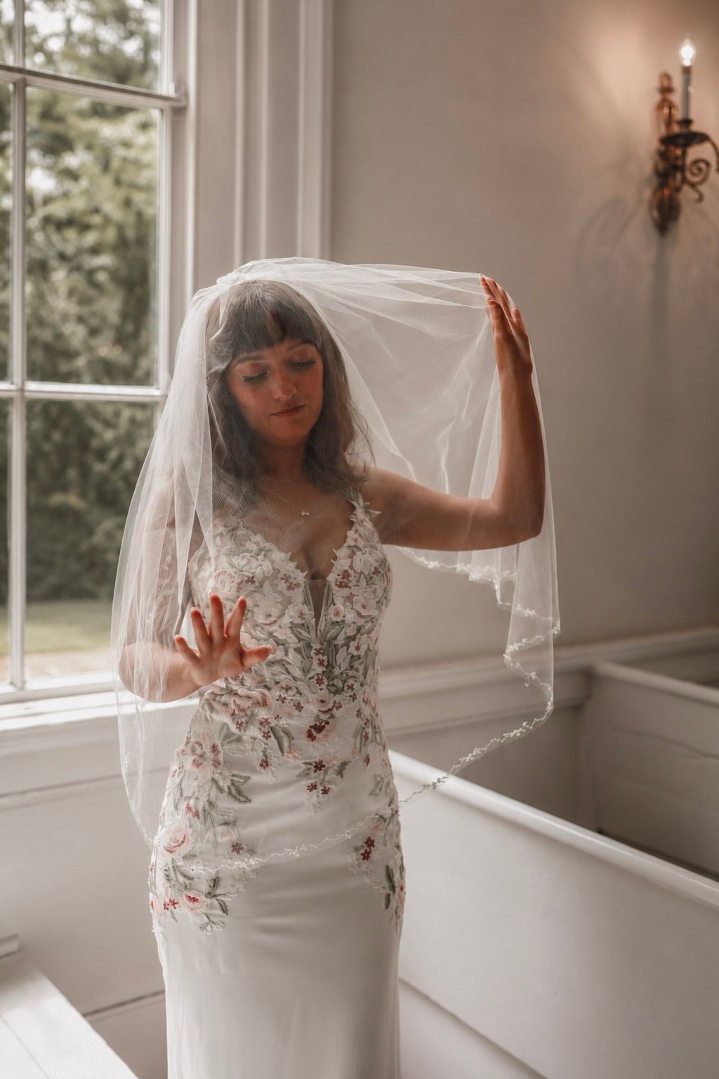 Bride in floral embroidered wedding dress lifting her veil near a bright window in an elegant indoor setting.