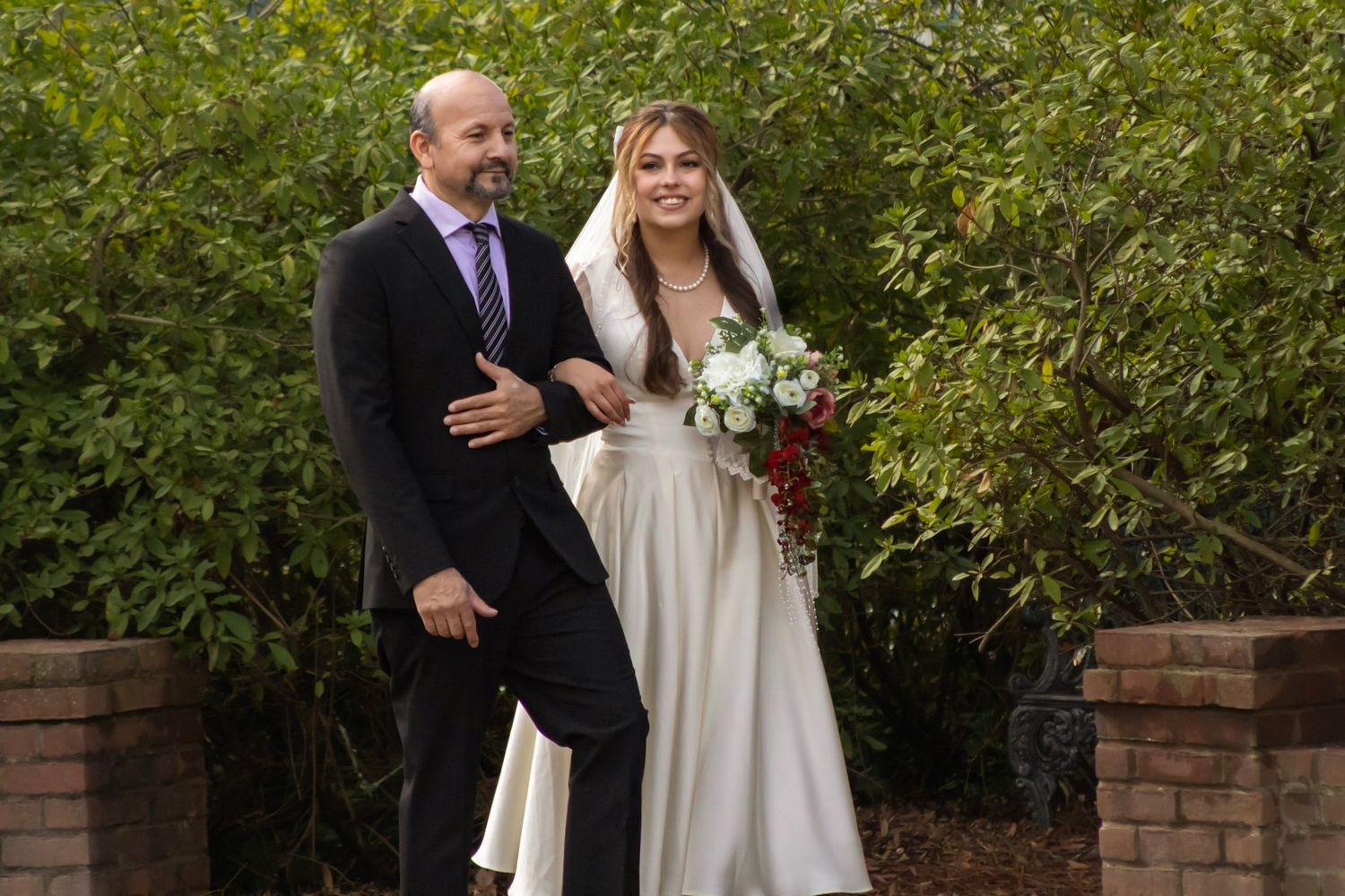Newlyweds embrace in outdoor garden ceremony with brick wall and natural foliage surroundings.