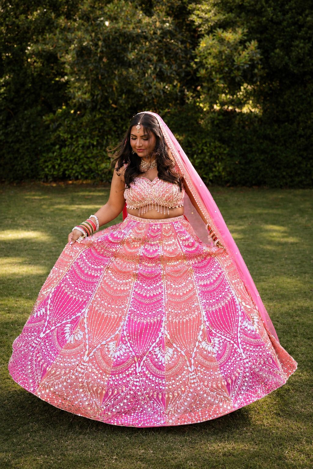 Woman in pink embroidered lehenga choli with dupatta twirling outdoors in garden setting.