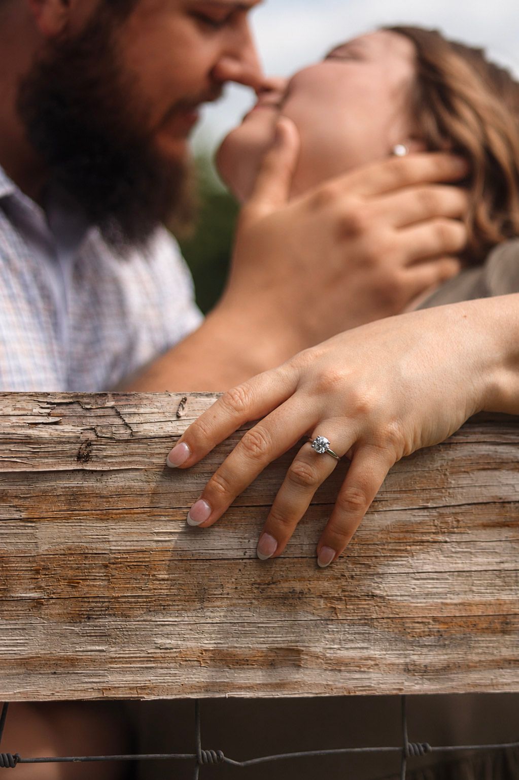 Couple sharing a kiss over a rustic wooden fence, with an engagement ring prominently displayed on the woman's hand.