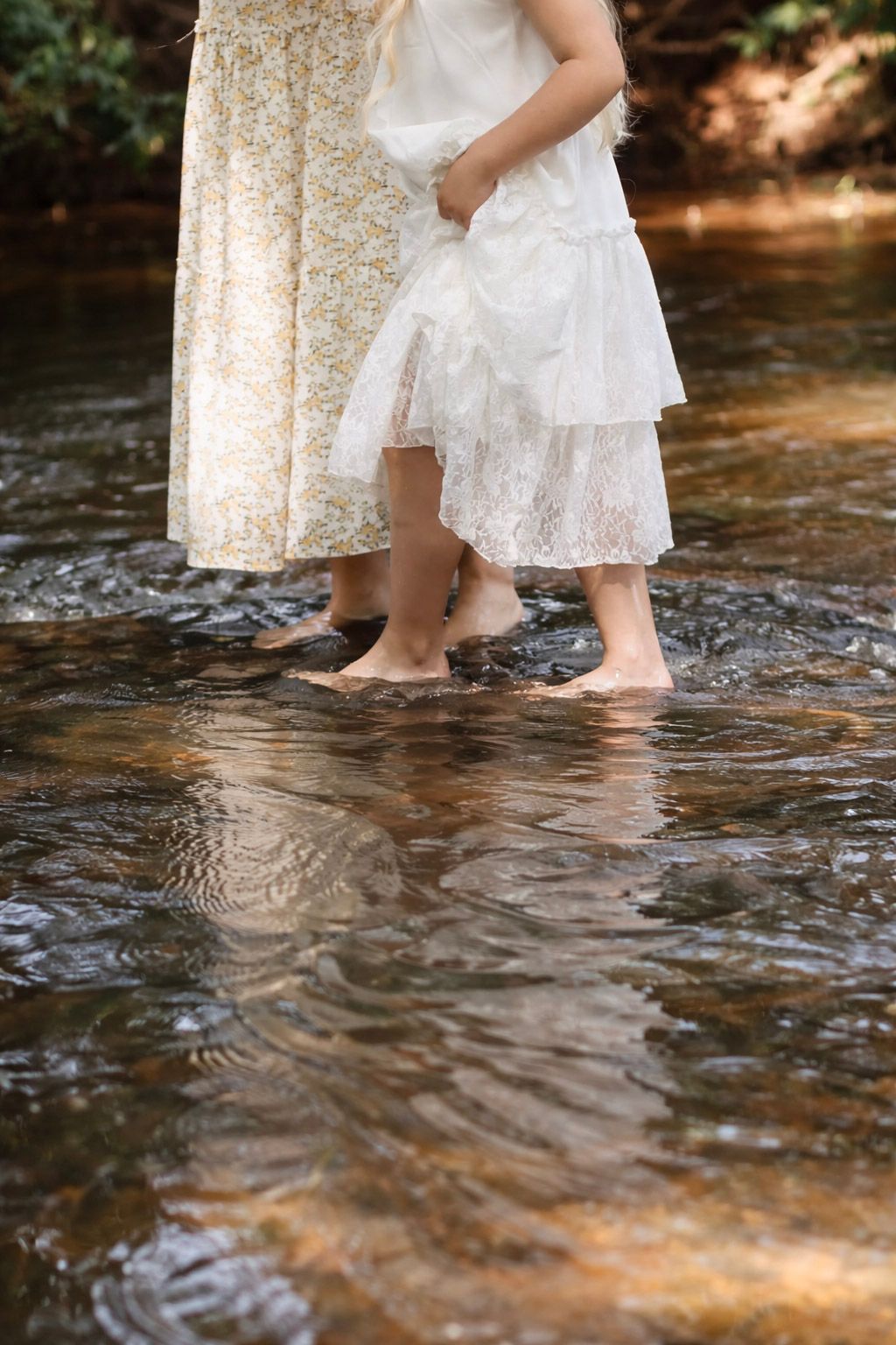 Two barefoot females in floral and lace dresses wading through a shallow rocky creek outdoors.