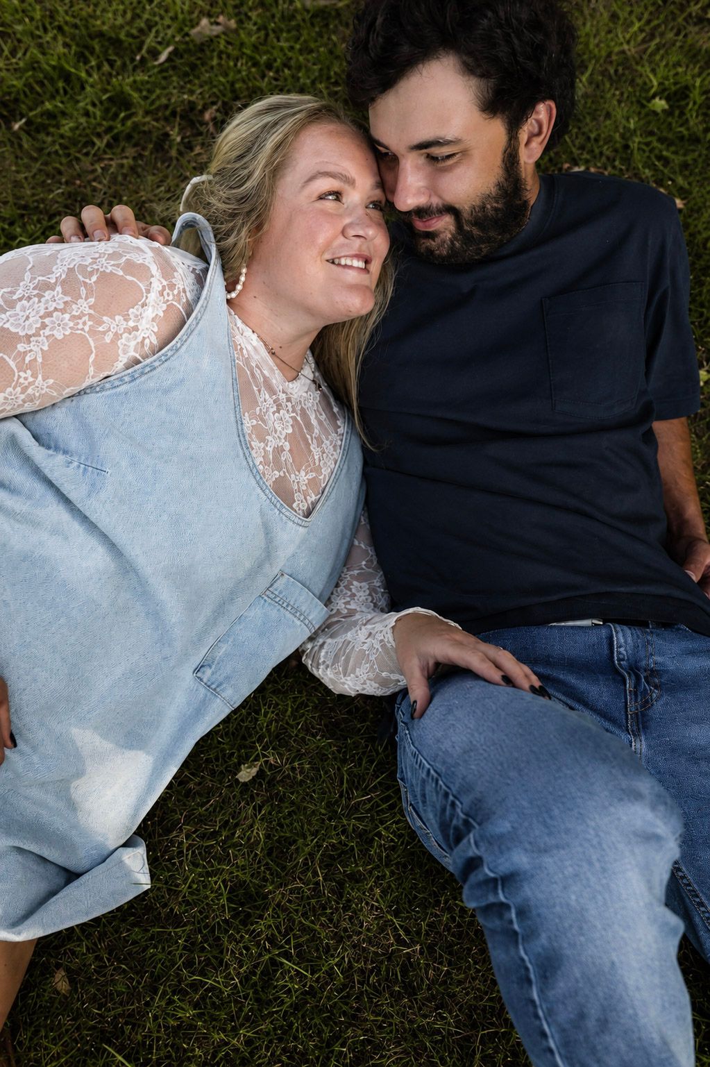 A smiling couple lying on green grass, gazing at each other, woman in denim overalls and lace top, man in navy shirt.