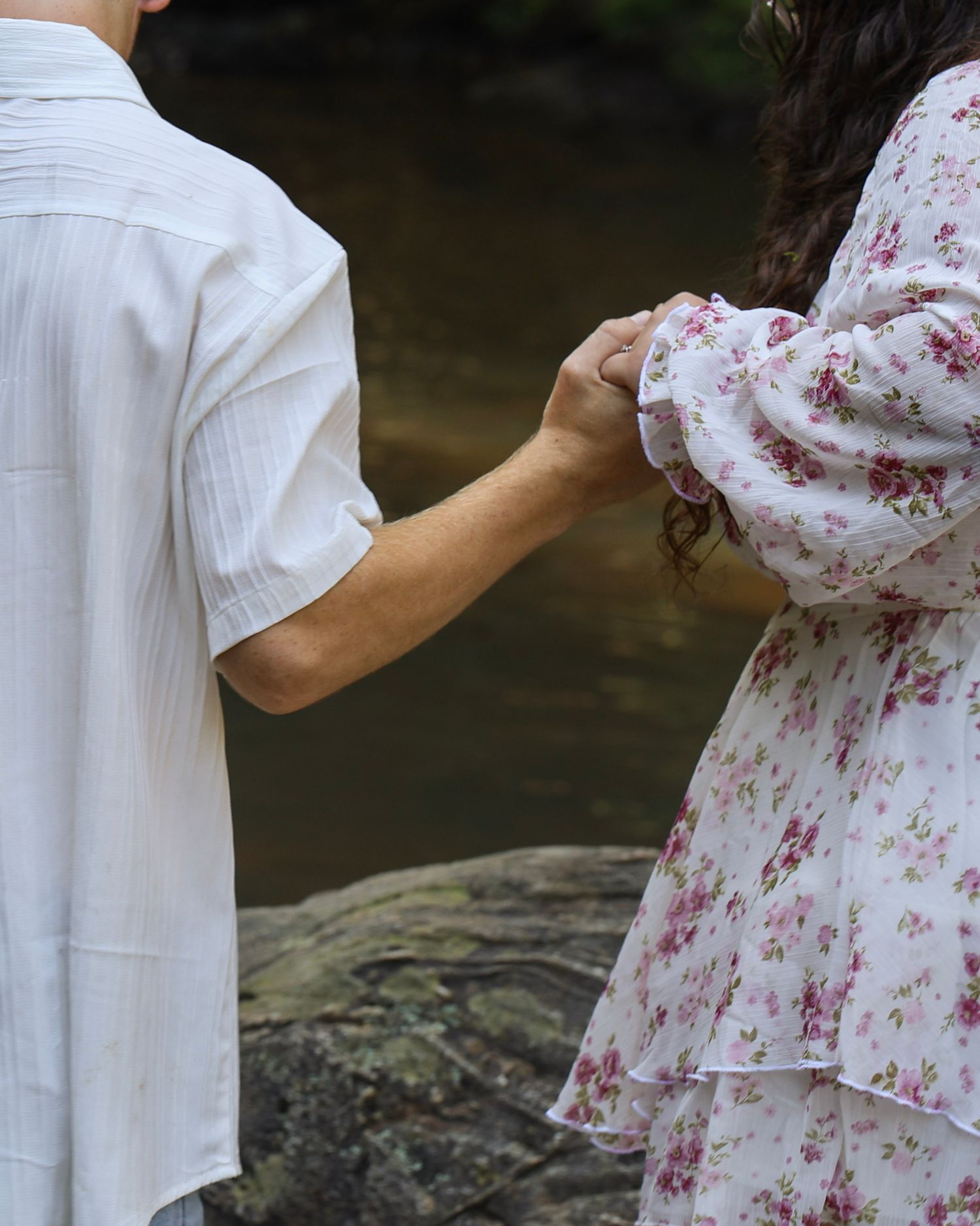 Couple holding hands outdoors near a rocky stream, wearing white shirt and floral dress.