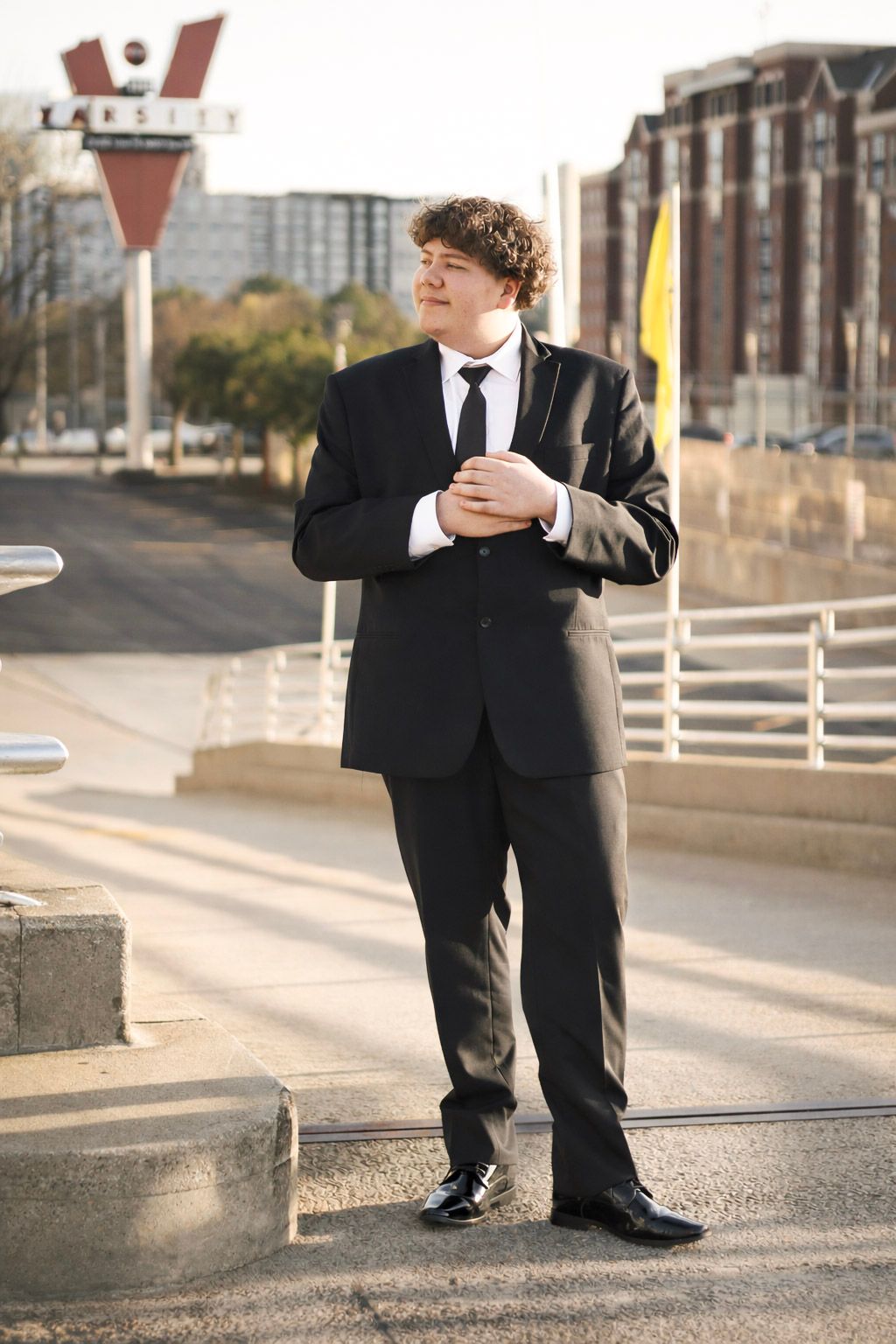 Young man in black suit and tie adjusting cufflinks outdoors near urban street with buildings in background.