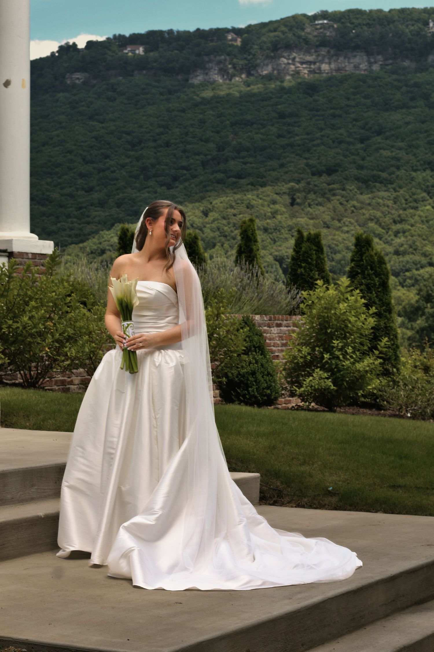 Elegant bride in white satin gown with veil holds bouquet on stone steps with lush mountain backdrop.