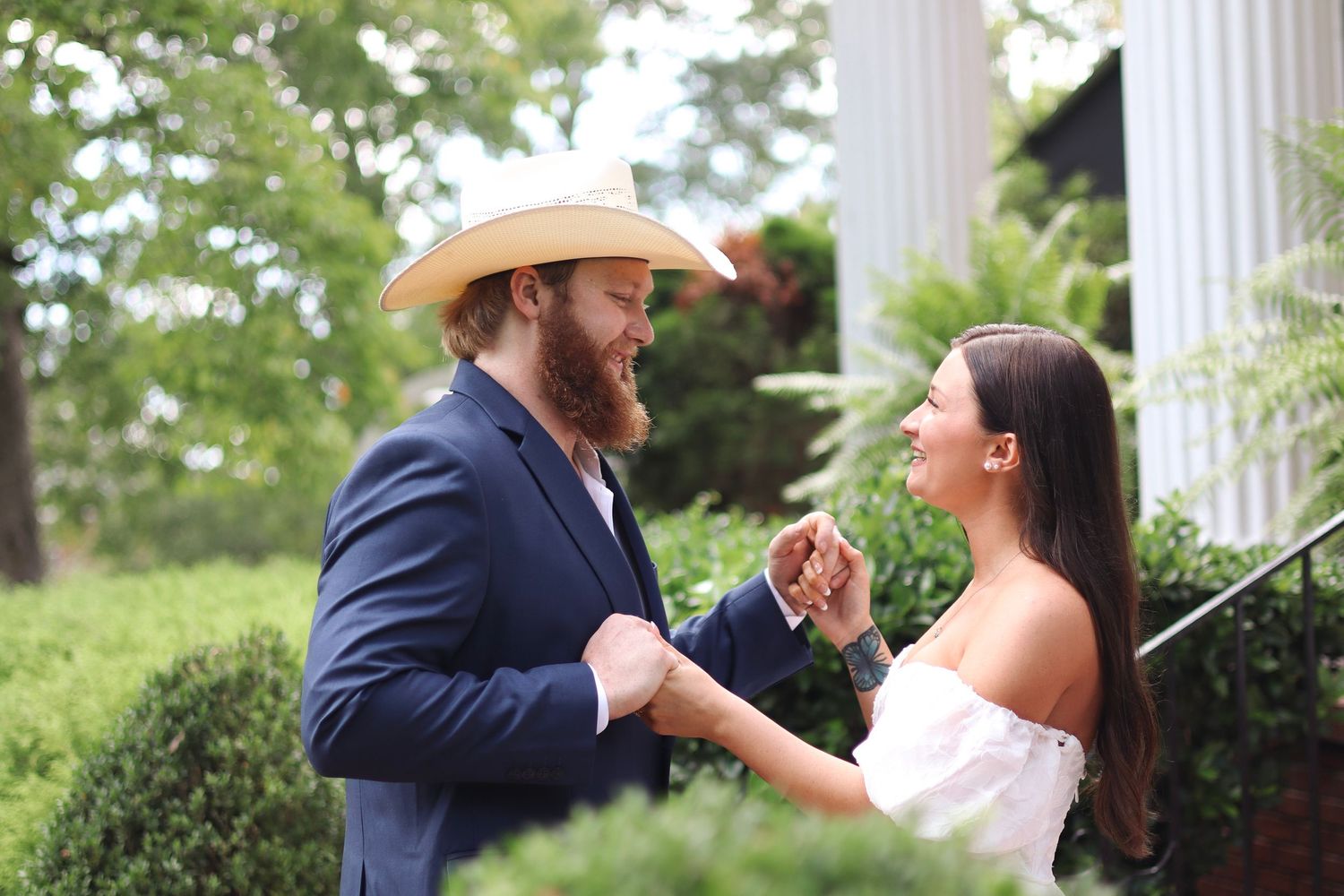 Two people share a candid moment outdoors, with white columns and greenery in the background.