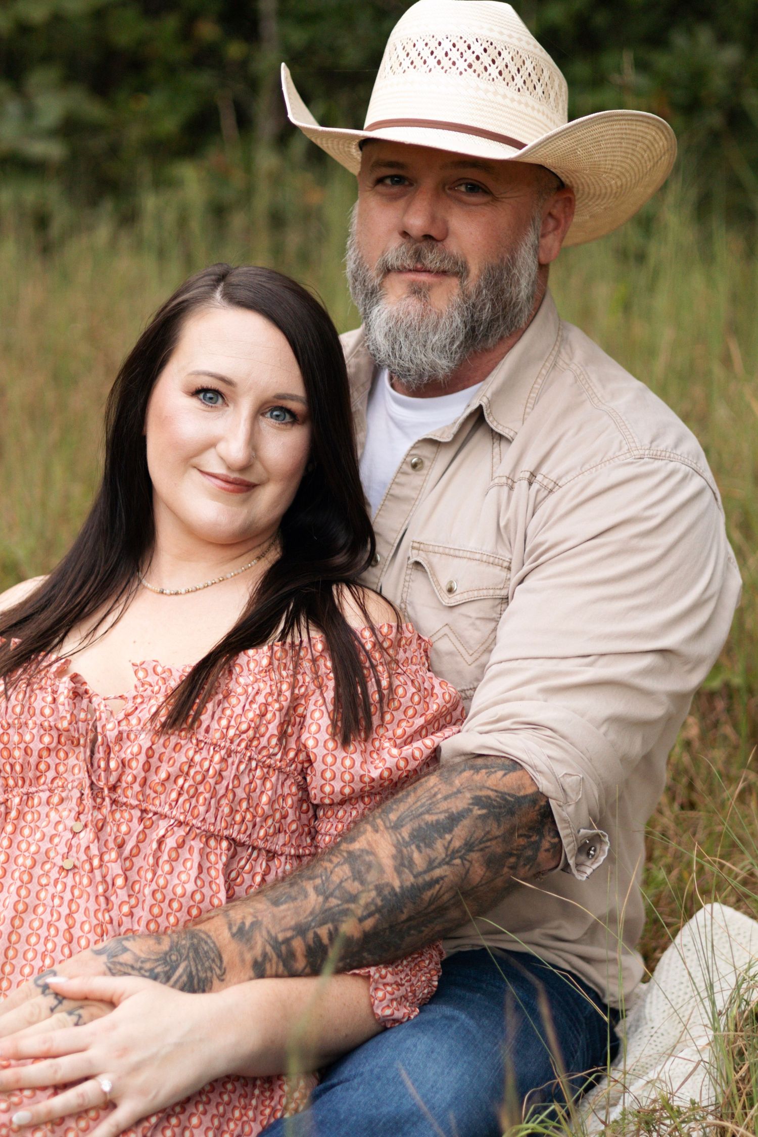 A couple in casual outdoor attire sits together in a grassy field.