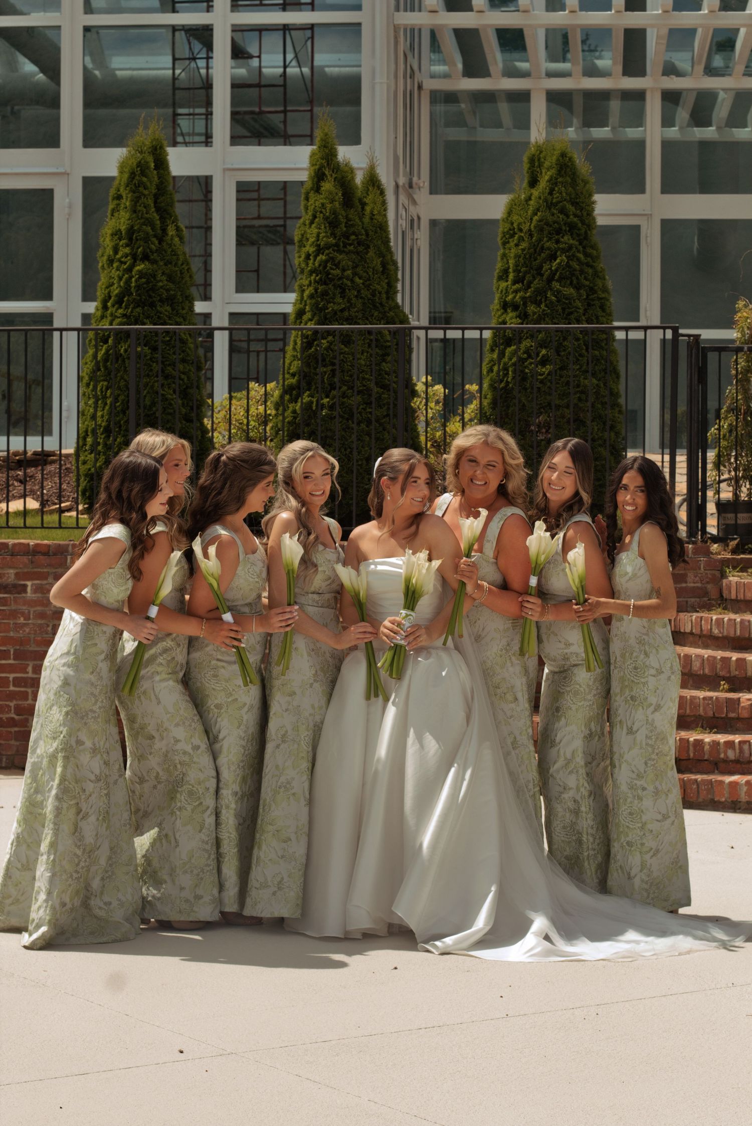 Bride in white gown surrounded by bridesmaids in sage green floral dresses holding white tulips outdoors.