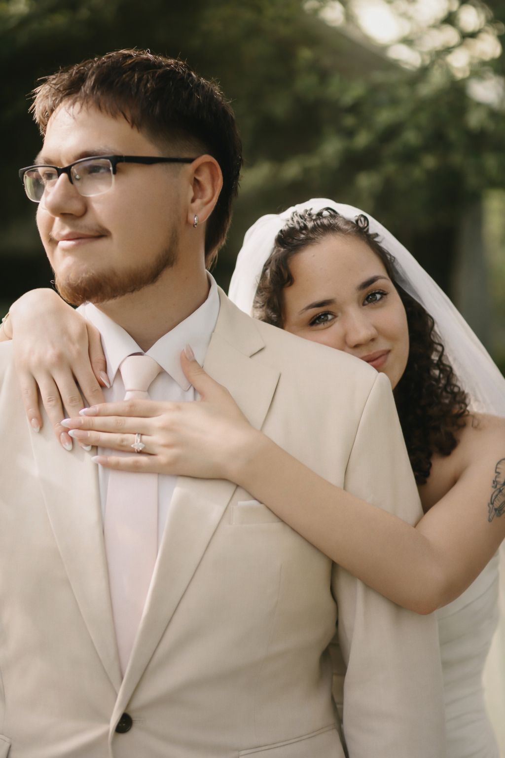 Bride in white veil embraces groom in beige suit from behind during outdoor wedding photo session.