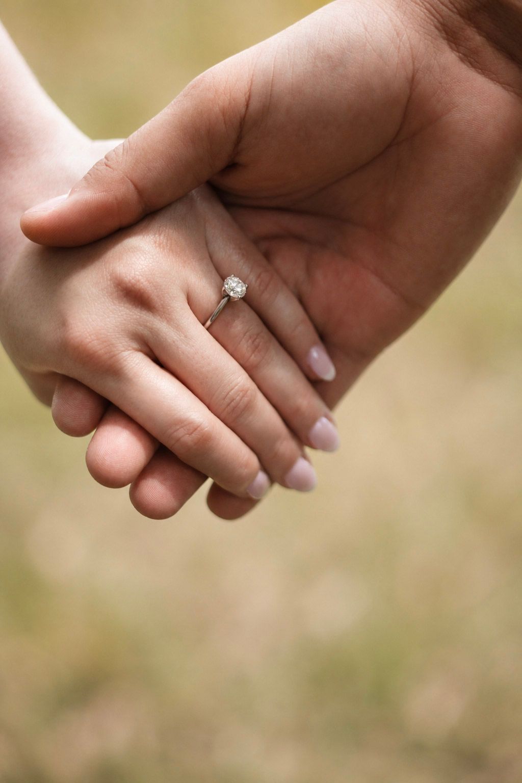 Two hands holding together, one wearing a sparkling diamond engagement ring with nude manicure, outdoors.
